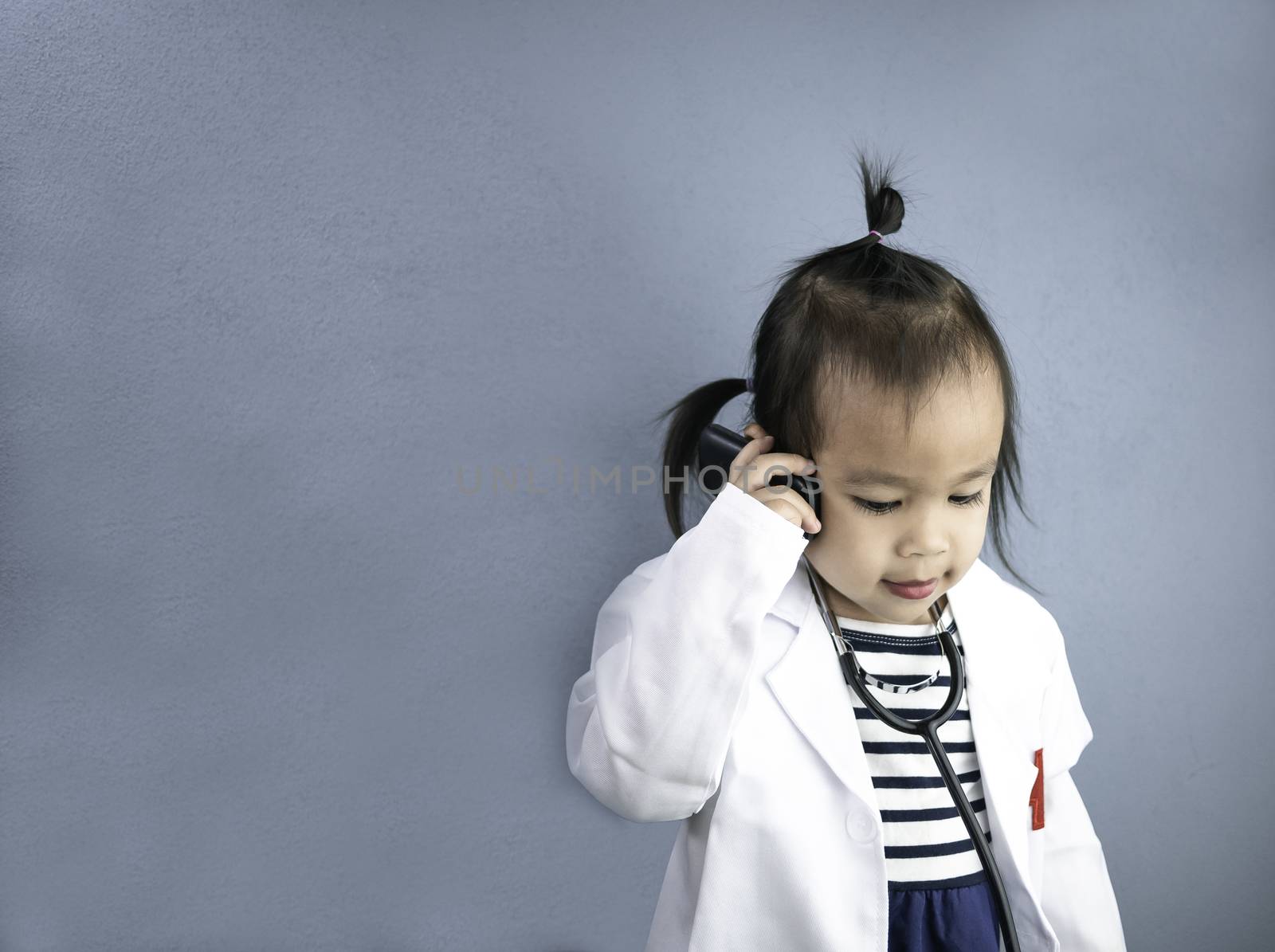 Asian little girl role playing doctor occupation wearing white gown uniform with smile and talking by phone. Playing is learning of children.