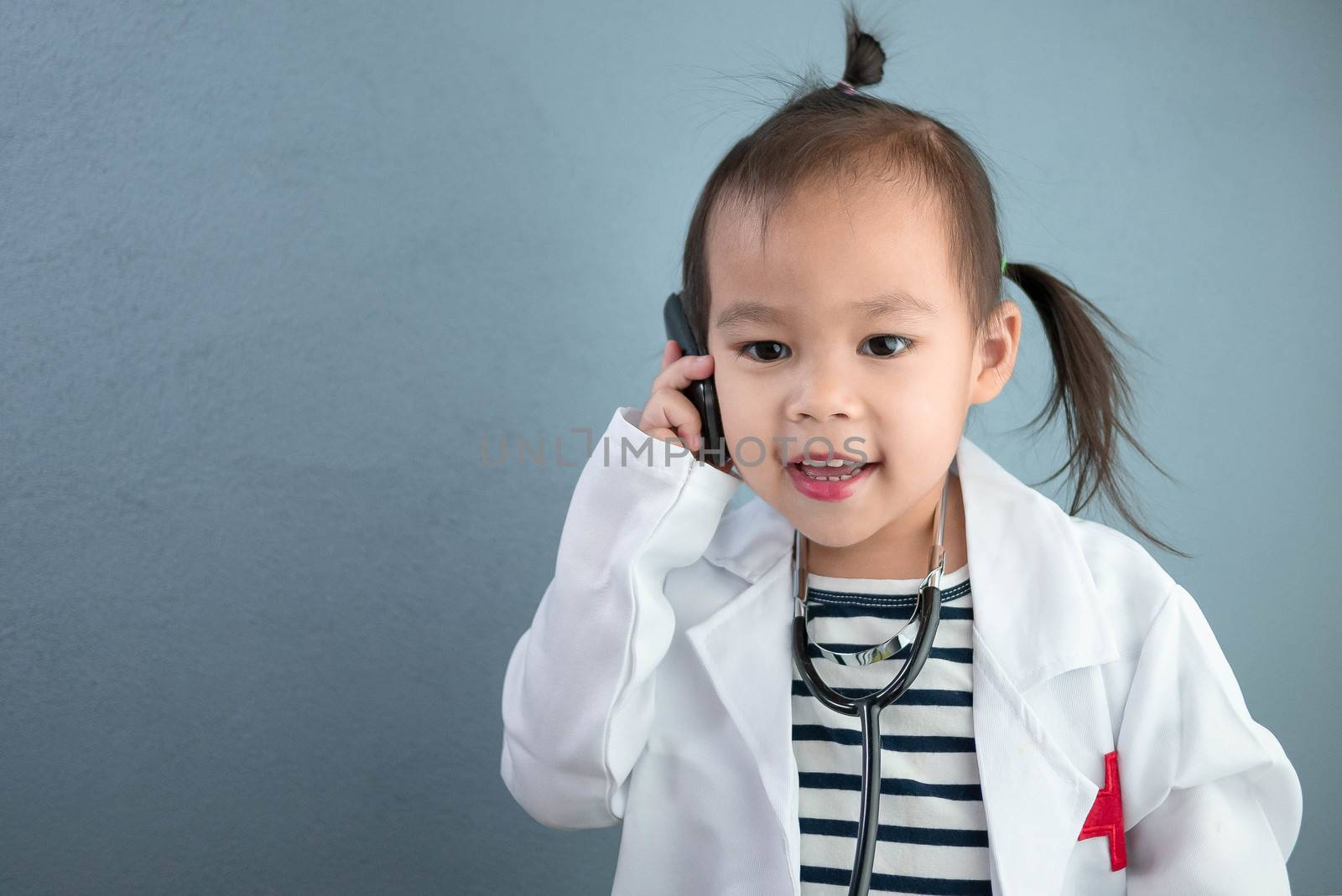 Asian little girl role playing doctor occupation wearing white gown uniform with smile and talking by phone. Playing is learning of children.