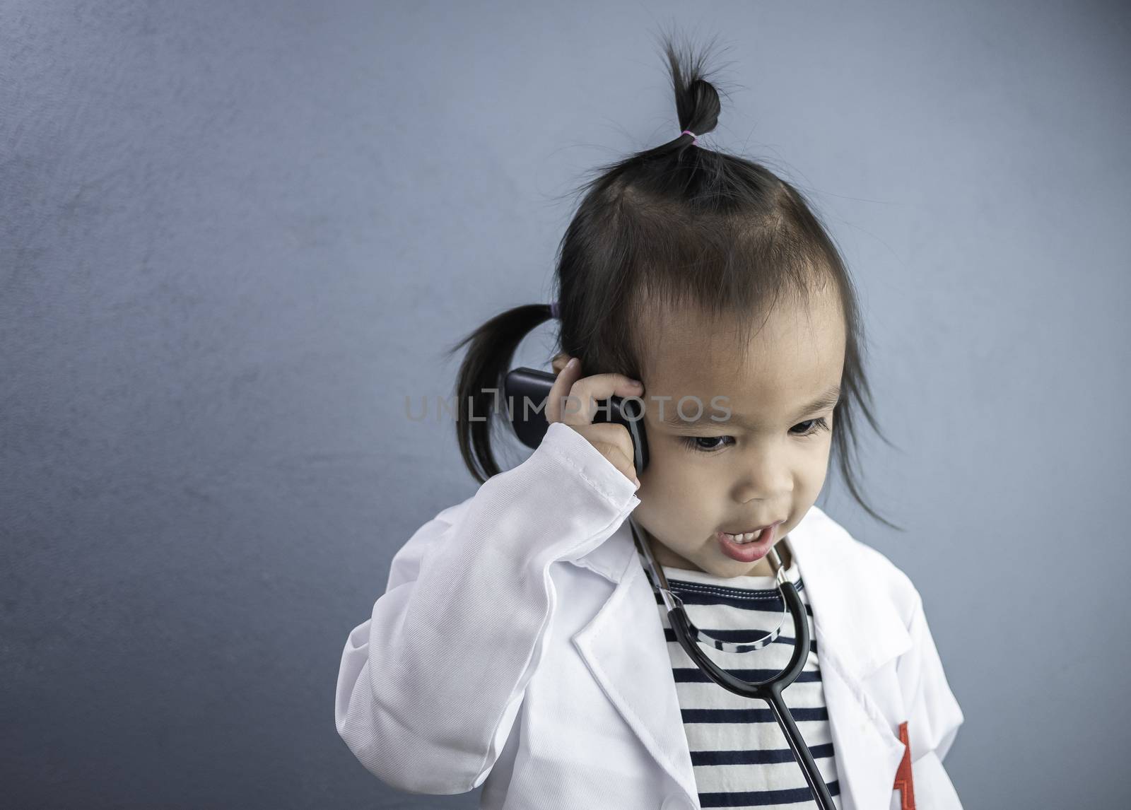 Asian little girl role playing doctor occupation wearing white gown uniform with smile and talking by phone. Playing is learning of children.