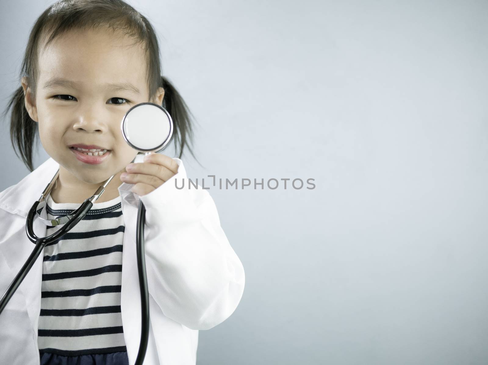 Asian little girl role playing doctor occupation wearing white gown uniform and holding a stethoscope. Playing is learning of children.