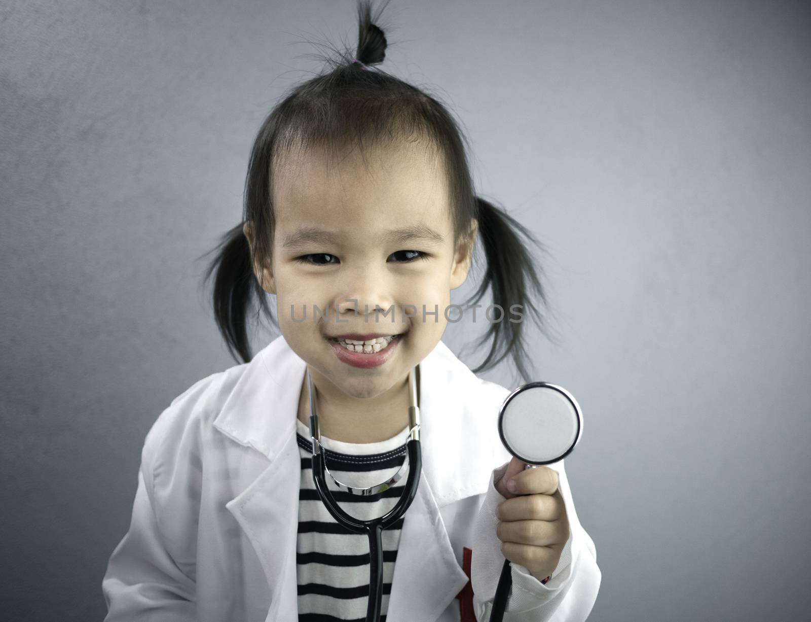 Asian little girl role playing doctor occupation wearing white gown uniform and holding a stethoscope. Playing is learning of children.