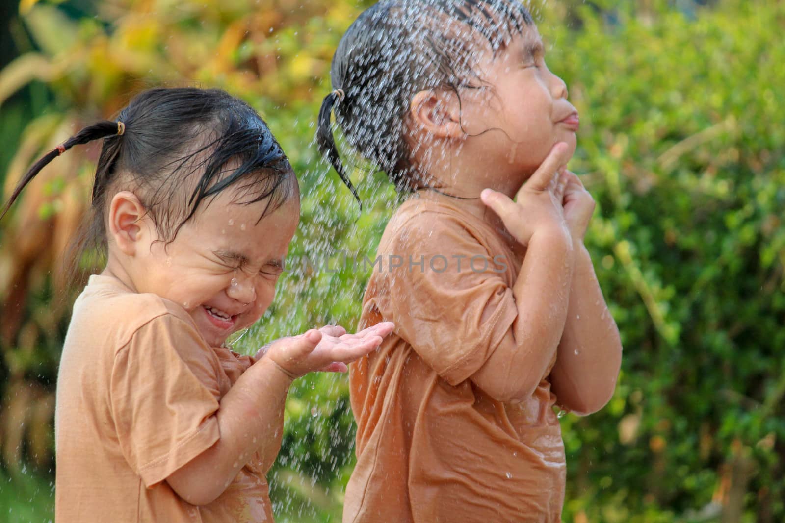 Cute asian little child girl ans her sister having fun to play with water spraying hose in summer garden.