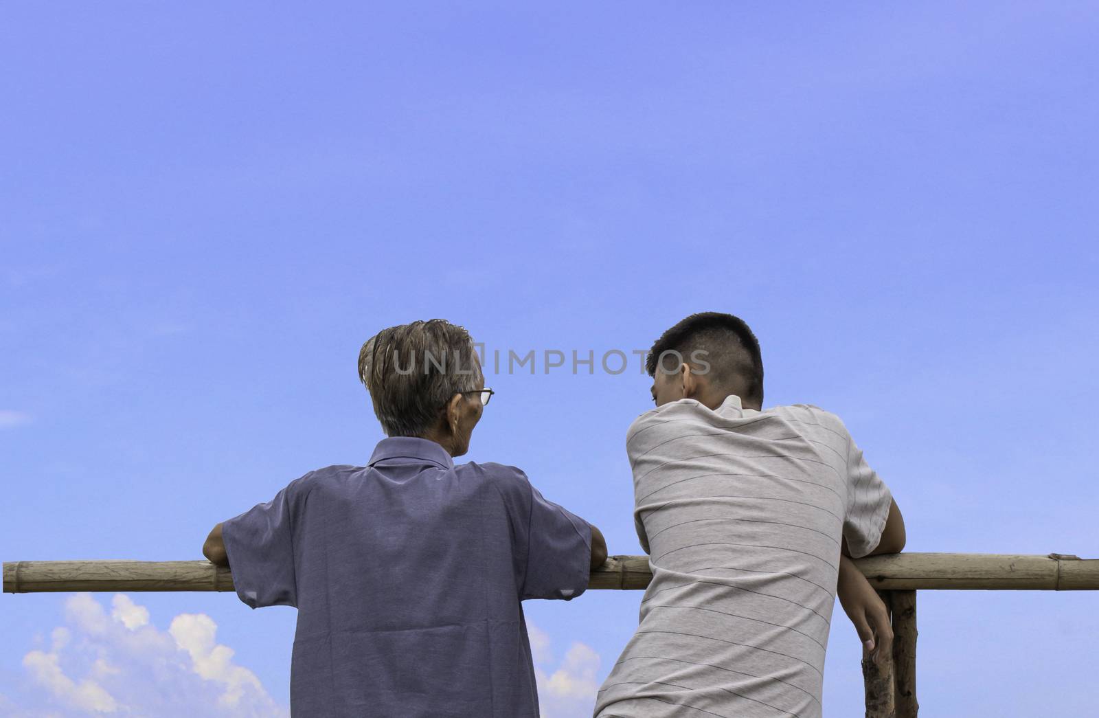 Rear of Asian teen boy and grandfather standing and talk while looking view on blue sky on mountain.