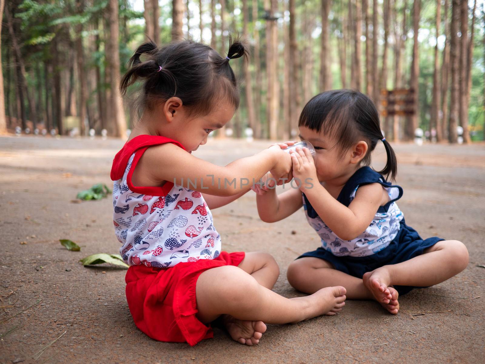 Asian little child sit in the garden and feed some milk to her sister from glass by TEERASAK