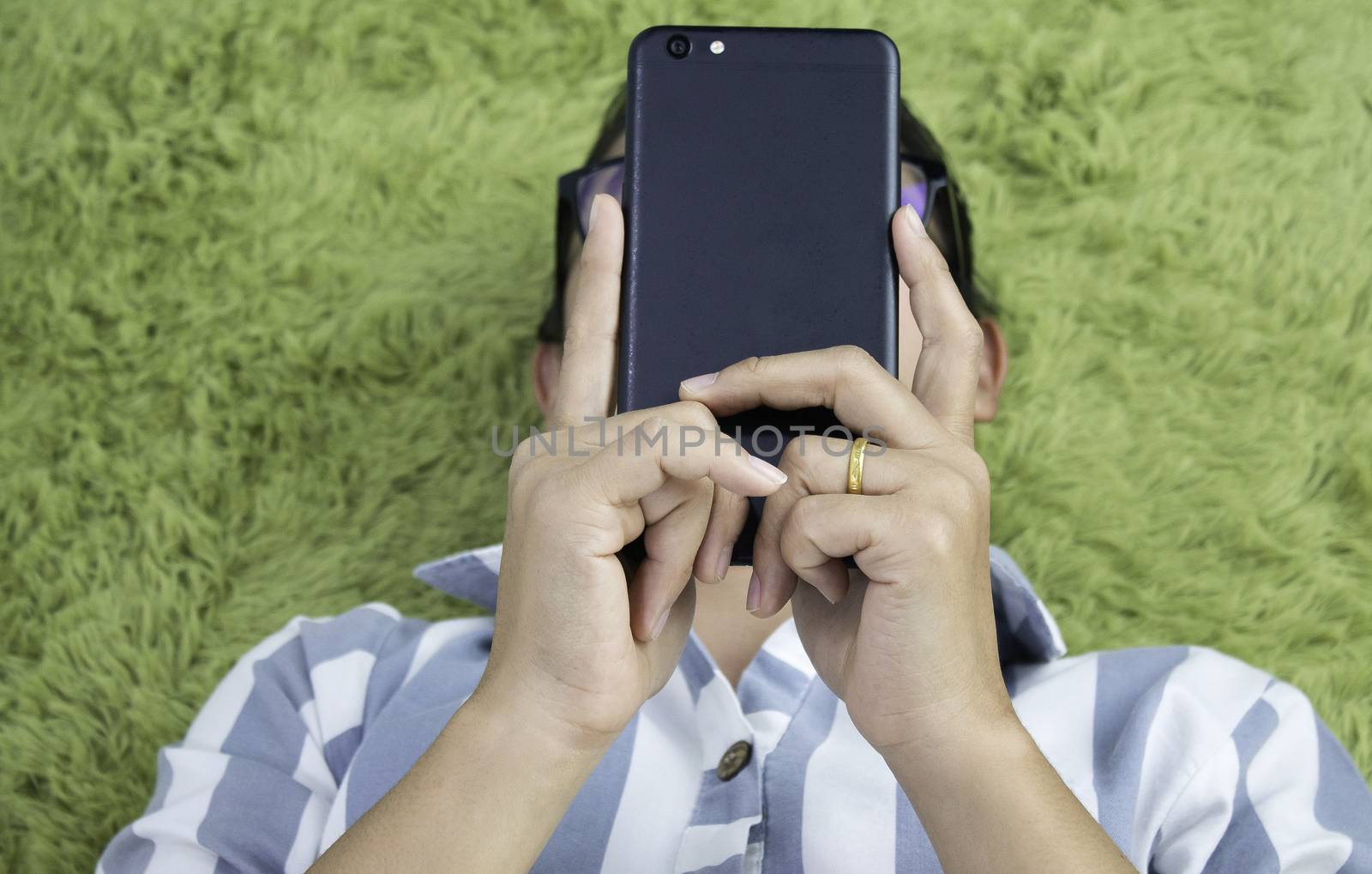 Close up of Asian young woman hand using a smartphone while lying on her back on green furry carpet at home. by TEERASAK