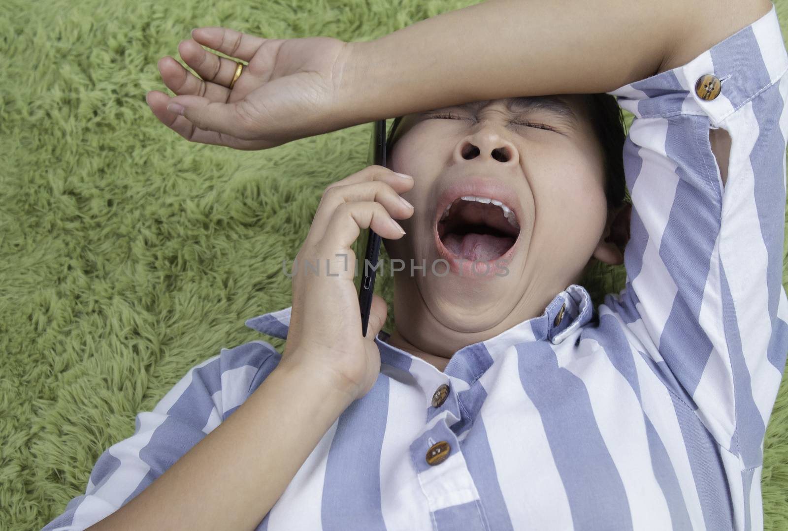 Close up of Asian young woman talk on smartphone while lying on her back and yawn on green furry carpet at home. by TEERASAK