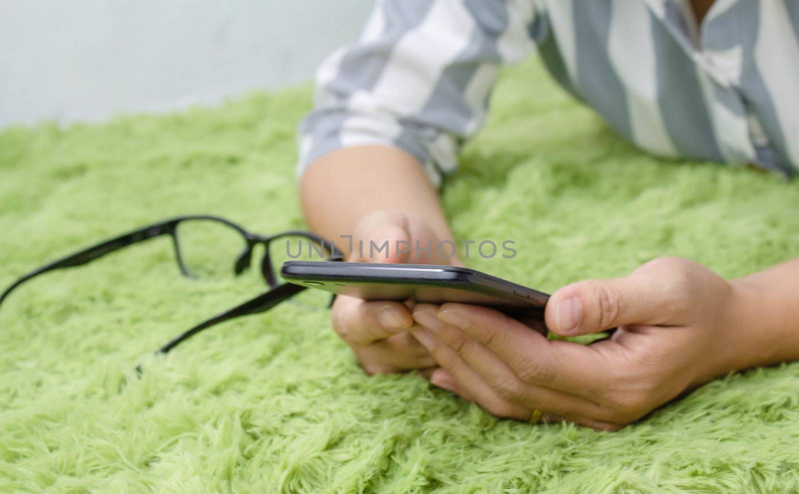 Close up of Asian young woman hand using a smartphone while lying down on green furry carpet at home. by TEERASAK