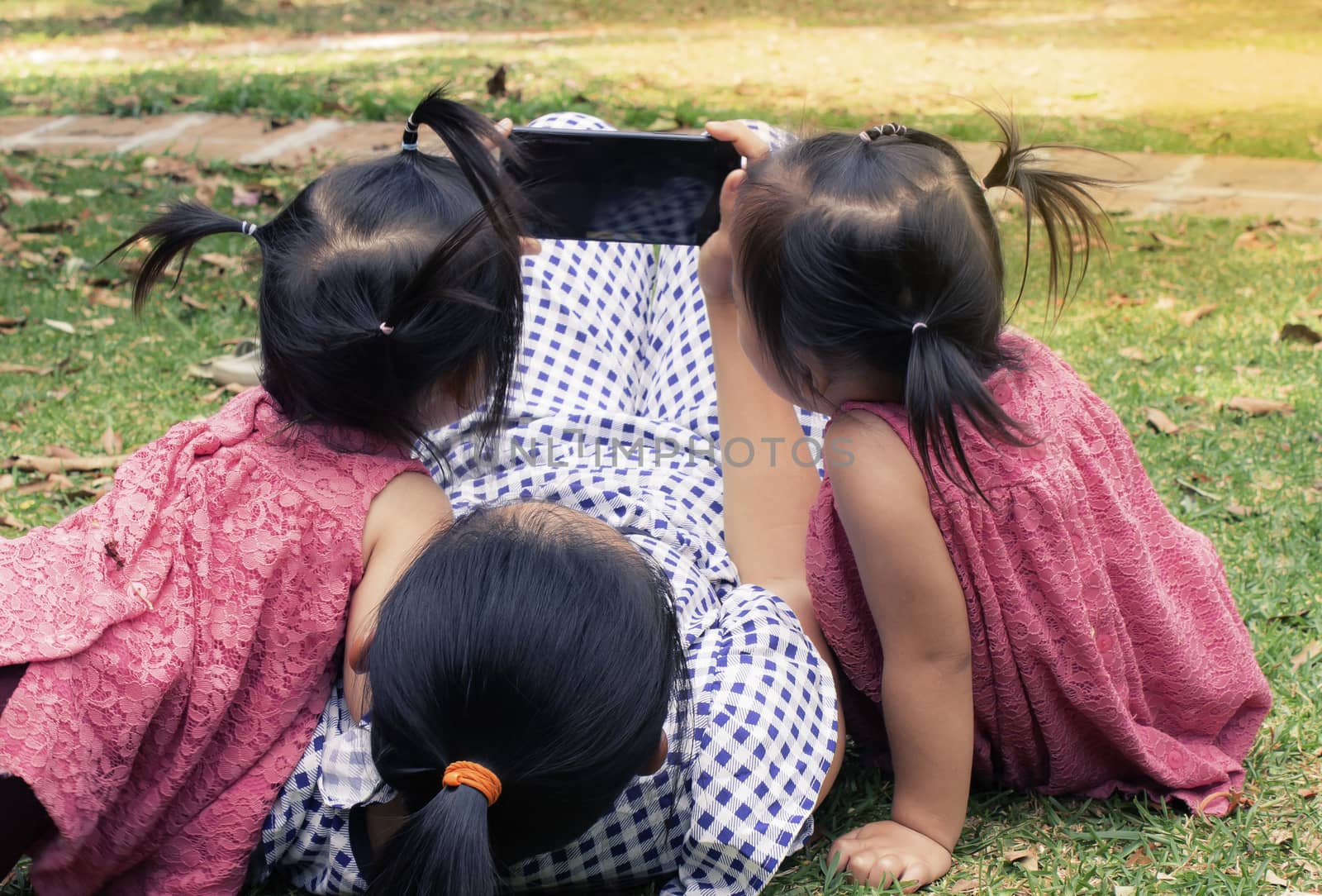 Asian mother and her daughters sitting on the grasses ground in the garden and looking at smartphone happily. by TEERASAK