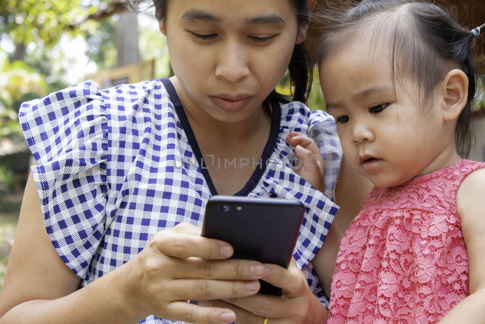 Asian mother and her daughter sitting on the grasses ground in the garden and looking at smartphone happily. by TEERASAK