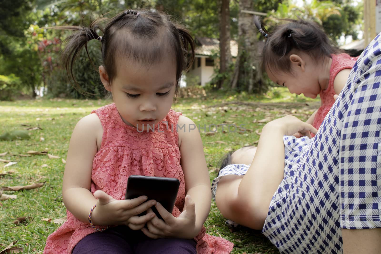 Asian little girl sitting on the grasses ground in the garden and looking at smartphone happily while mother and sister lying down to play  near. by TEERASAK