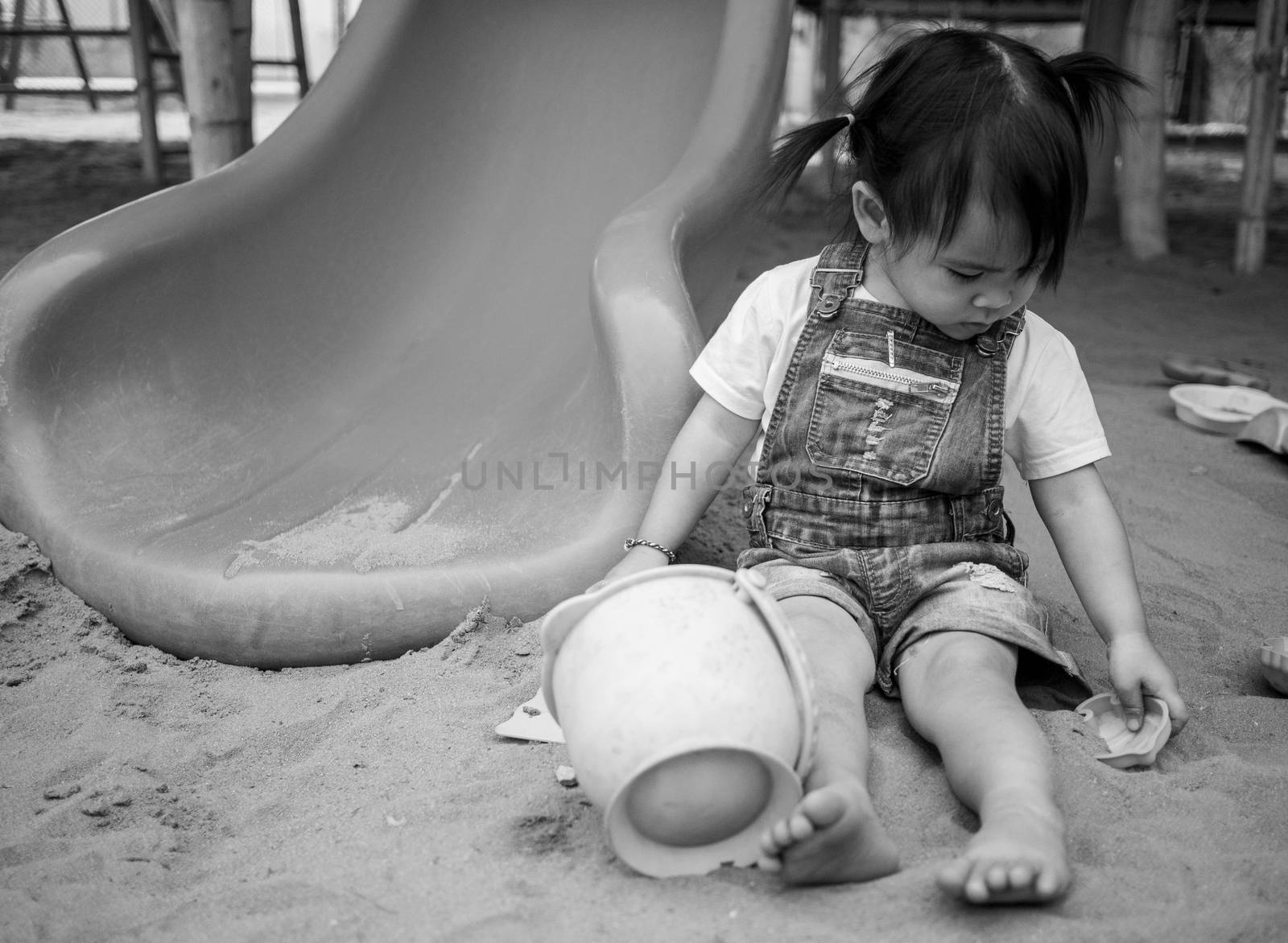 Asian little girl sitting on sand ground in the playground and play a sand with plastic beach toys happily. Playing is learning for children. Black-white picture. by TEERASAK