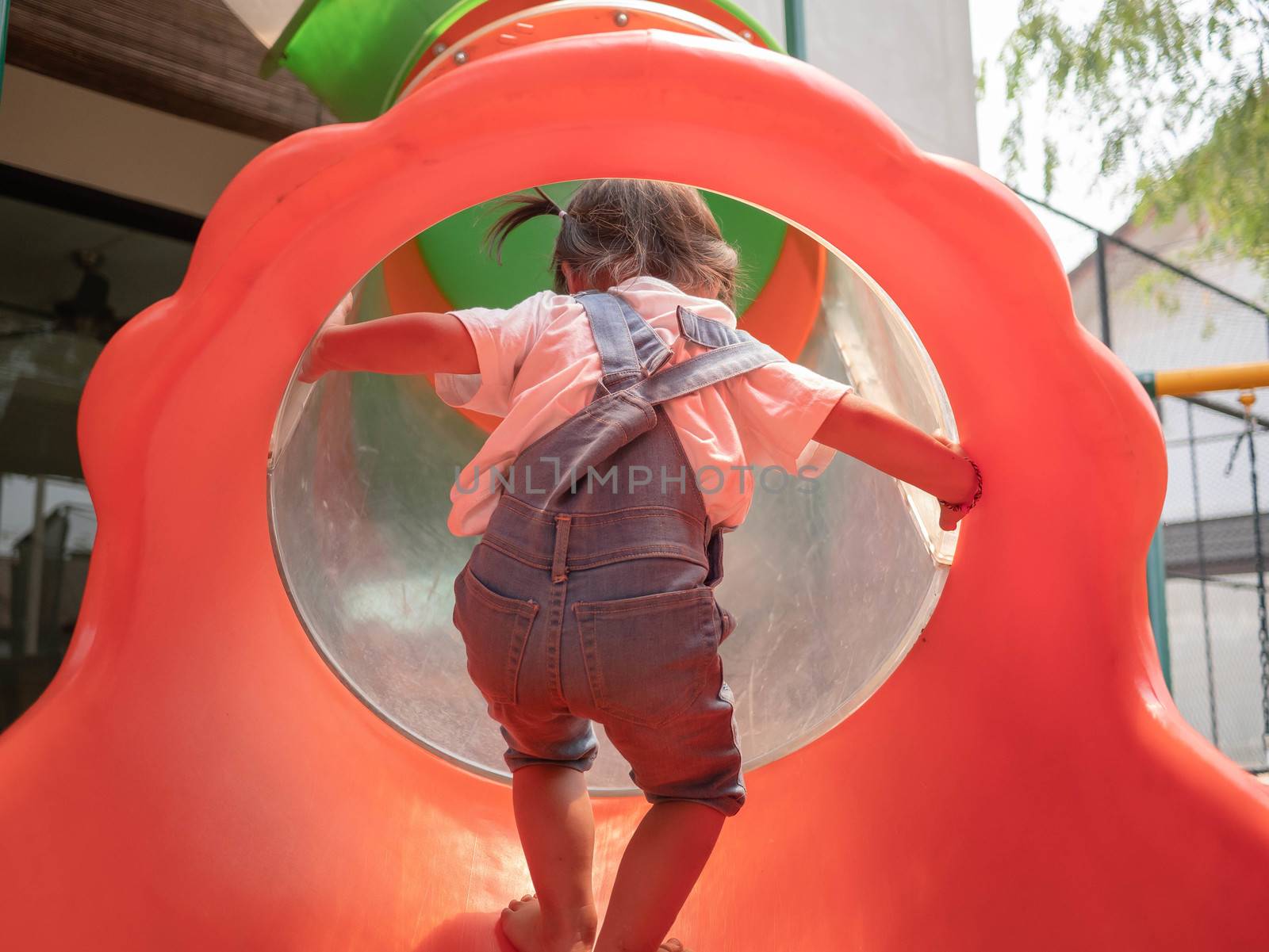 Asian little girl Tried to climb into the red tunnel tube slide at park playground. Playing is learning for children. Dangerous in children concept. by TEERASAK