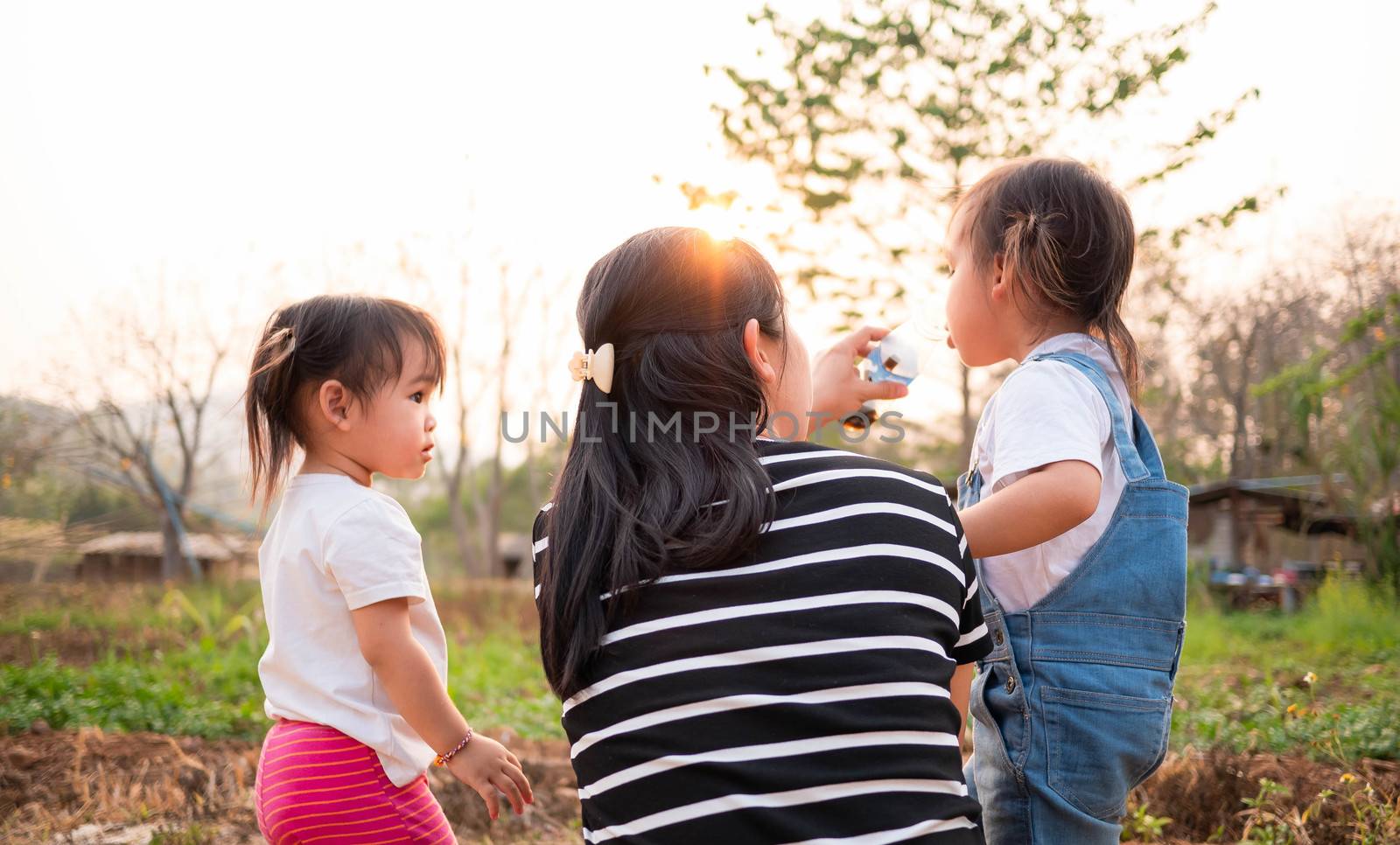 Asian little child girl drink water from bottle after tired of playing in the garden with mother and sister, looking sunset in evening. by TEERASAK