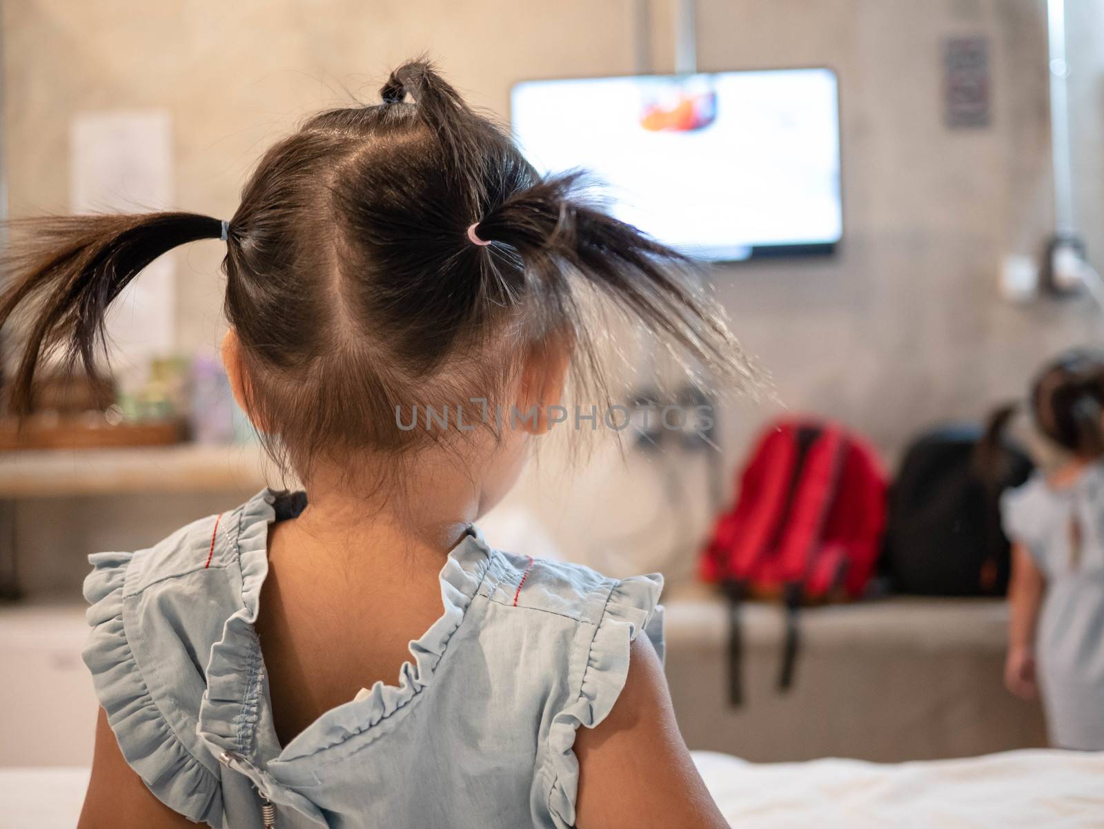 Back view of Asian little child girl sitting on the bed and looking television in the room.