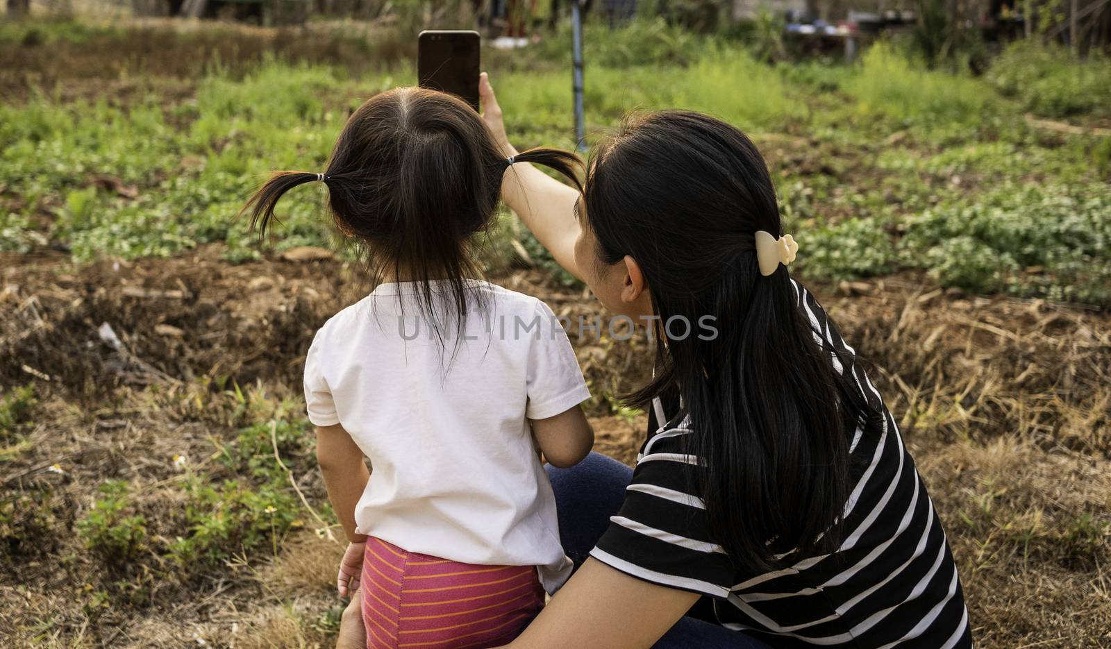 Back view of Asian little child girl taking a photo with mother in garden over sunset background.