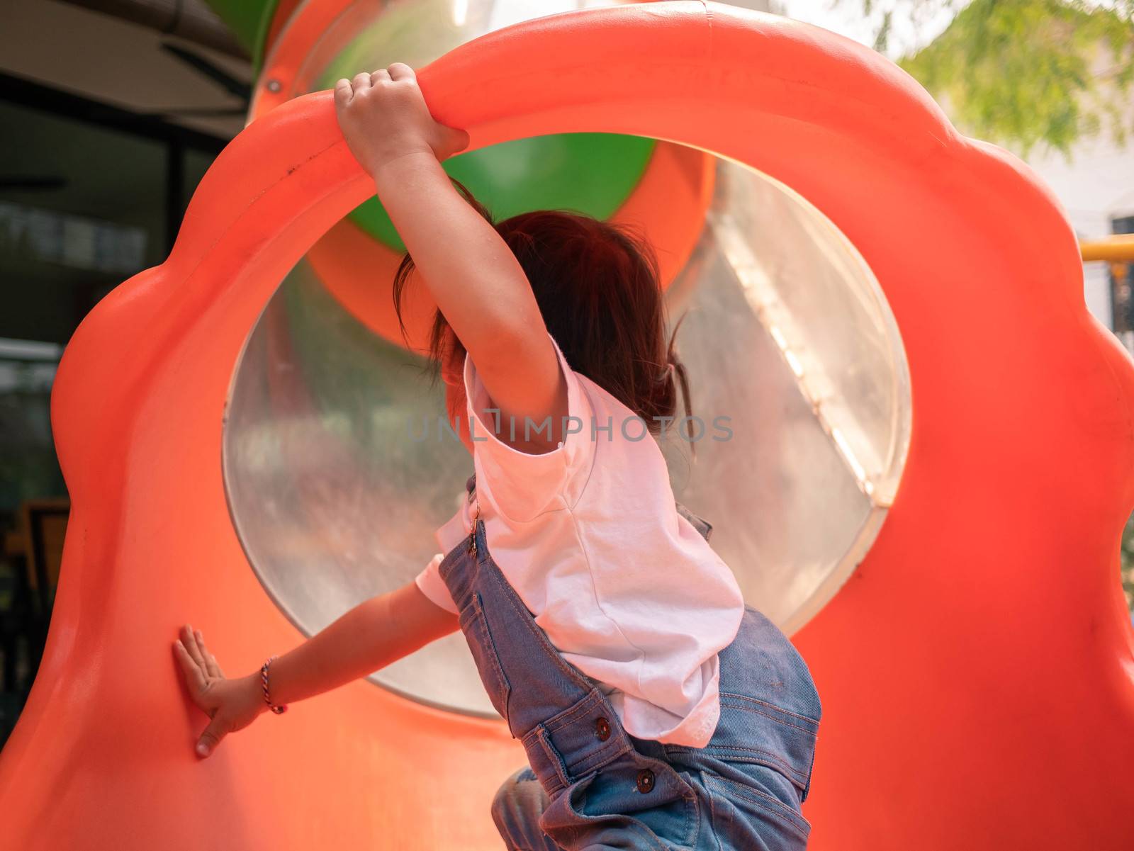 Asian little girl Tried to climb into the red tunnel tube slide at park playground. Playing is learning for children. Dangerous in children concept. by TEERASAK