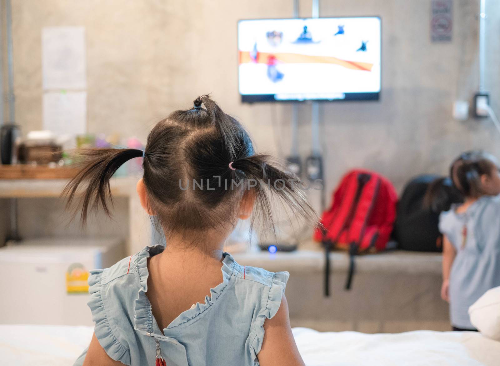 Back view of Asian little child girl sitting on the bed and looking television in the room. by TEERASAK