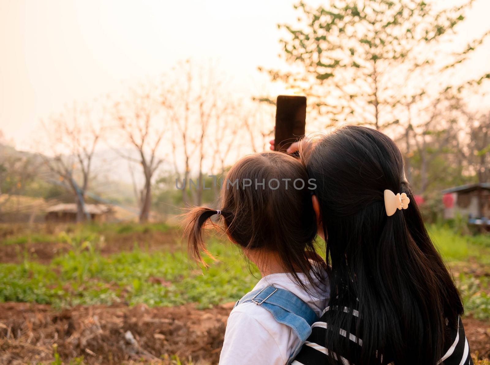Back view of Asian little child girl taking a photo with mother in garden over sunset background. by TEERASAK