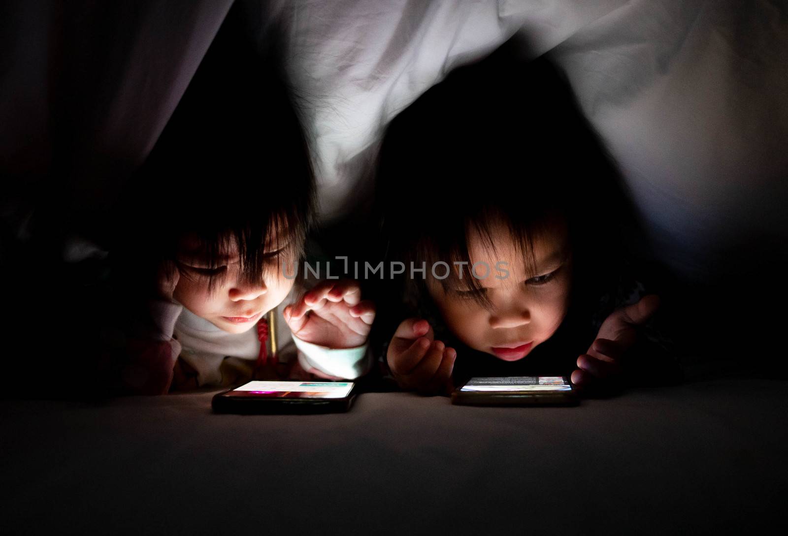 Portrait of Asian little girl with sister look cartoon on a smartphone under a blanket on the bed. The child's face is illuminated by a bright monitor. Health care concept.