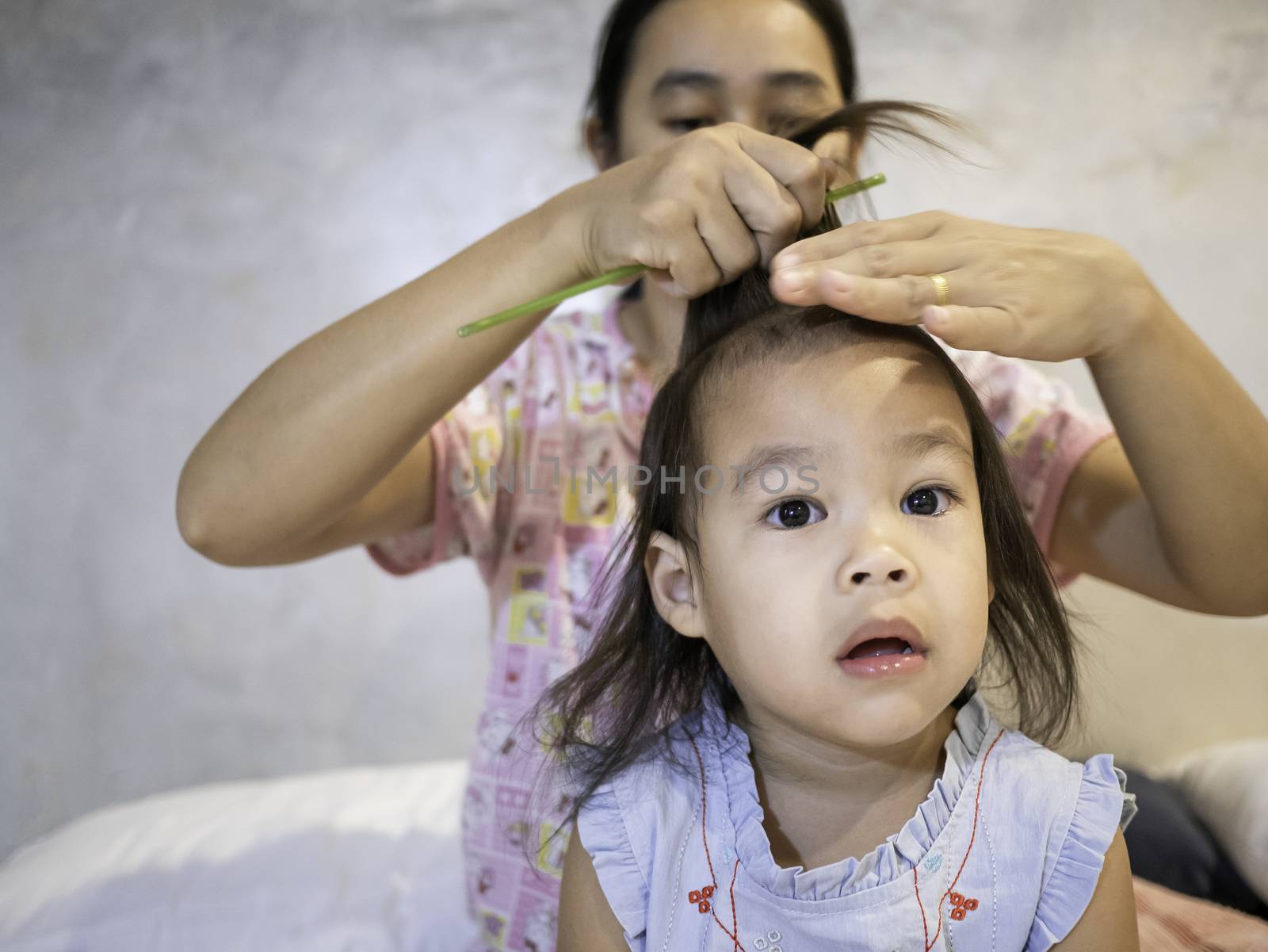 Charming Asian little girl is looking at camera and smiling while her young mother is combing daughter's hair at morning in the room.