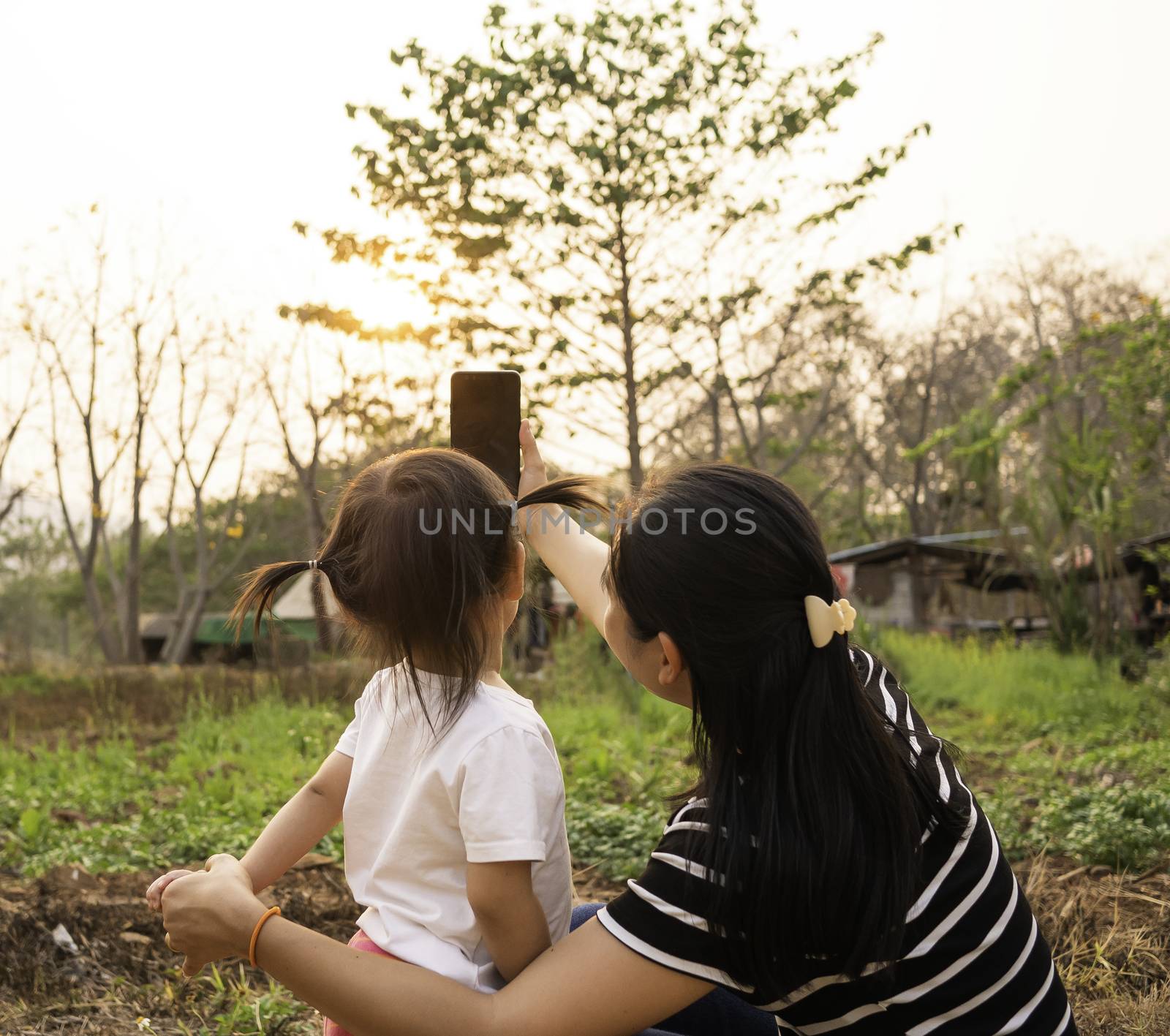 Back view of Asian little child girl taking a photo with mother in garden over sunset background. by TEERASAK