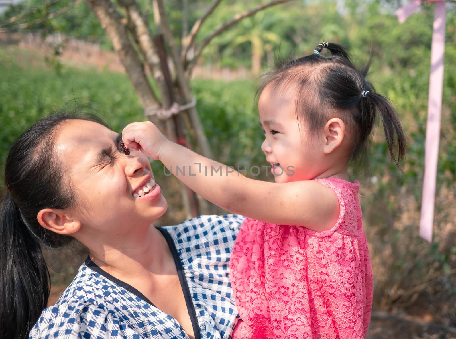Asian little girl make funny face to her mom; Pretend mother while mom holding her and walking in the garden. by TEERASAK
