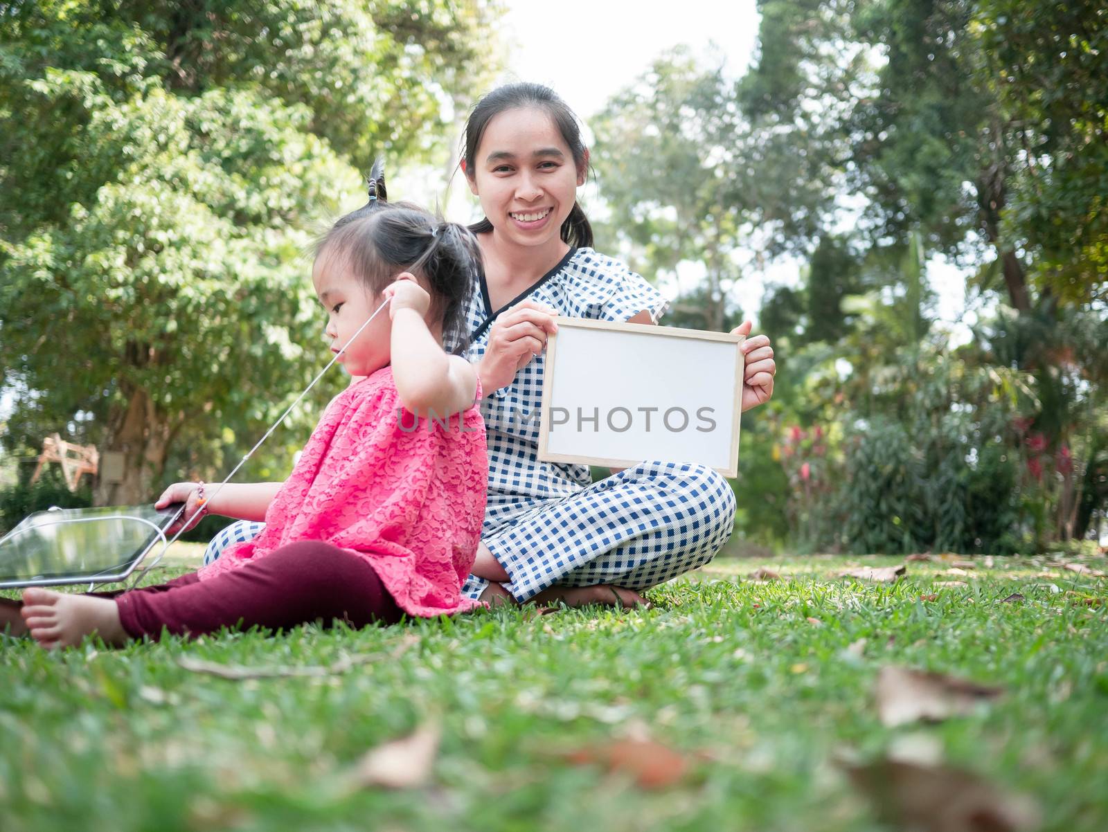 Asian young woman holding blank whiteboard on head, pointing finger and sitting on grass ground in the park. Space for your text.