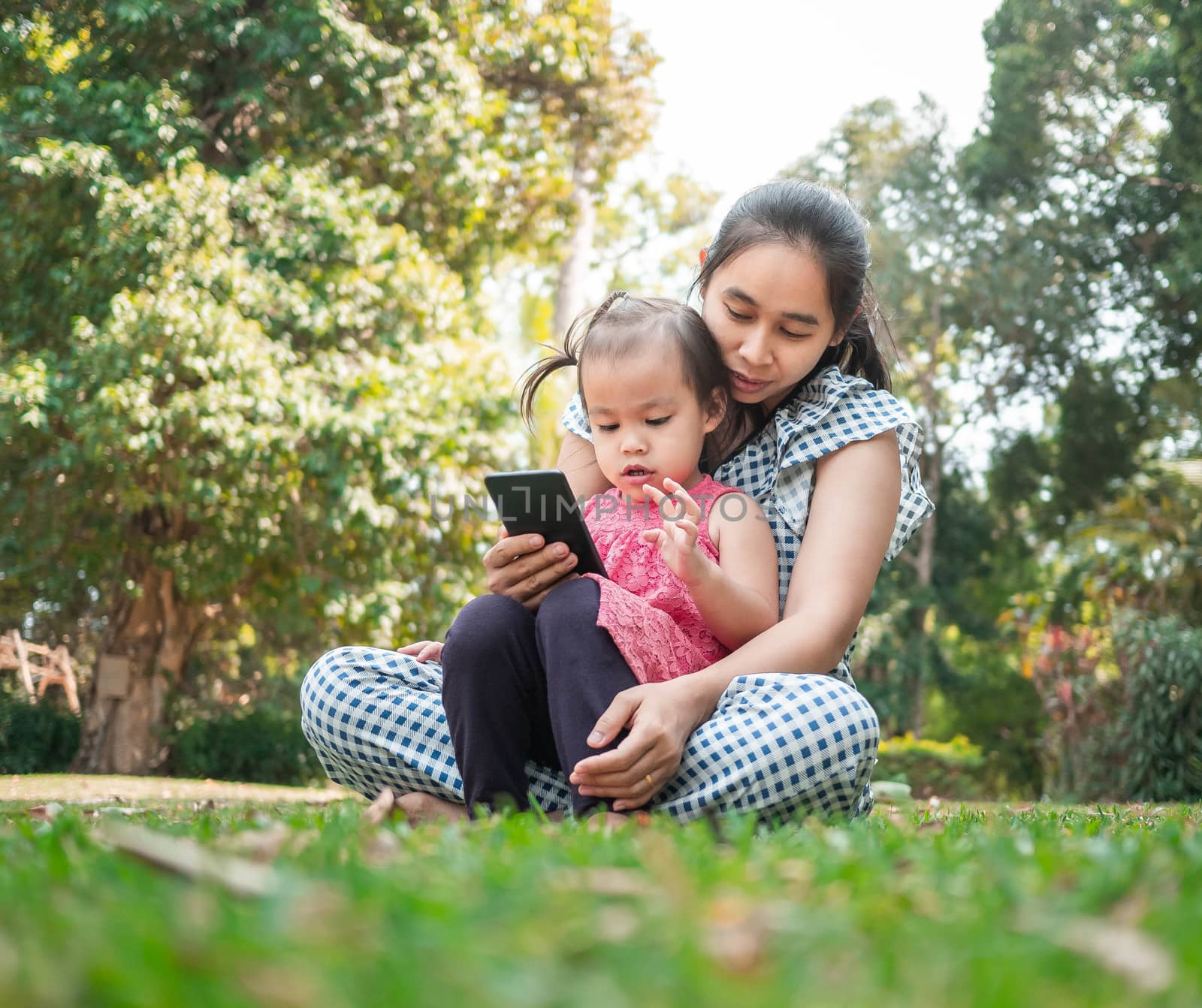 Asian mother and her daughter sitting on the grasses ground in the garden and looking at smartphone happily. by TEERASAK