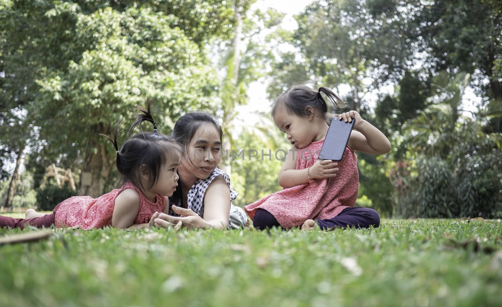 Asian mother and her daughters sitting on the grasses ground in the garden and looking at digital tablet happily. by TEERASAK