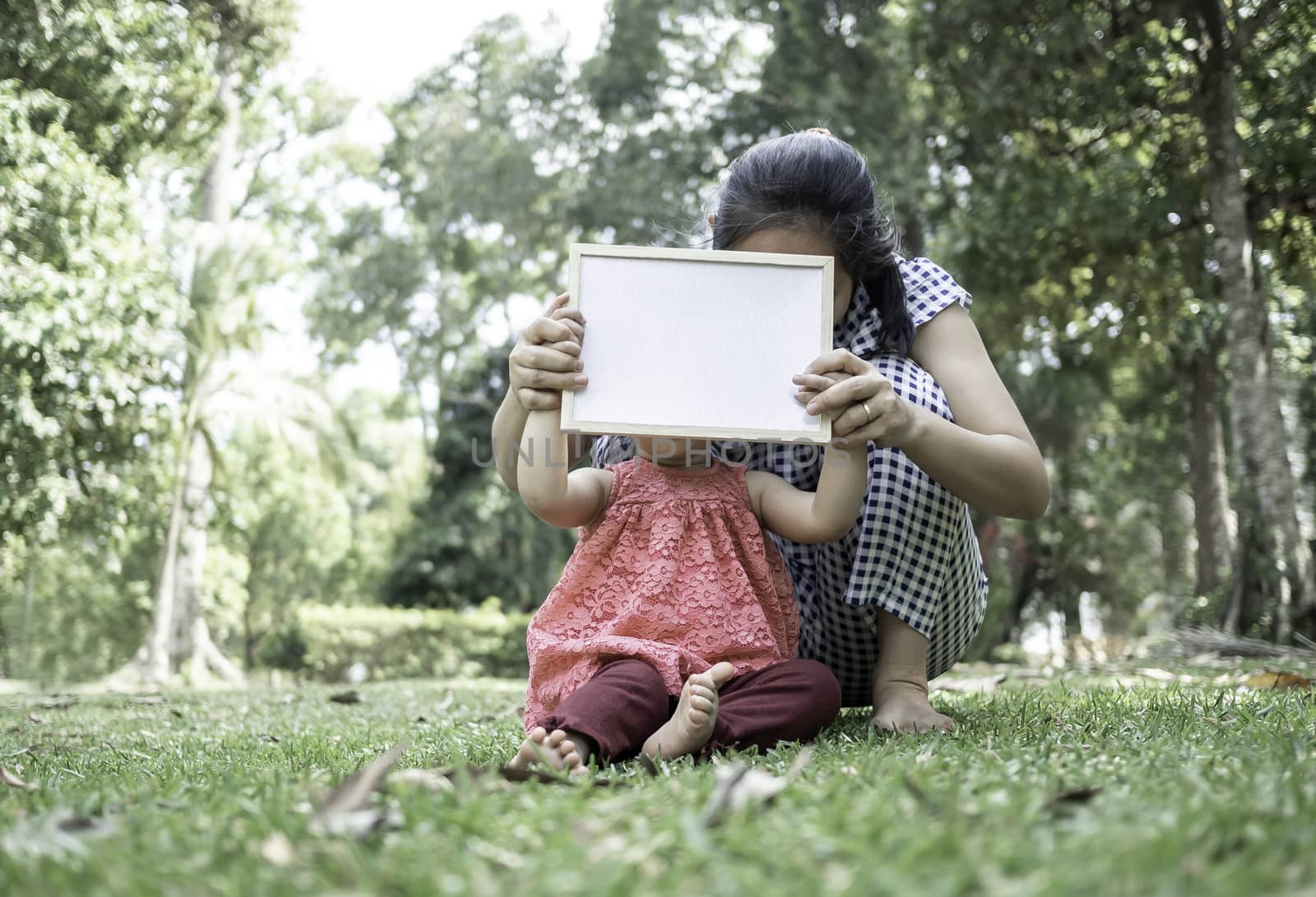 Asian little girl holding empty white board with mom and sitting on grasses ground. Space for write your text. by TEERASAK