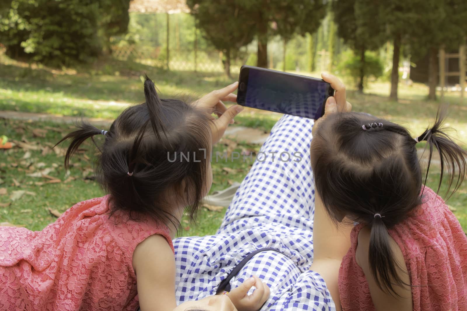 Asian mother and her daughters sitting on the grasses ground in the garden and looking at smartphone happily.