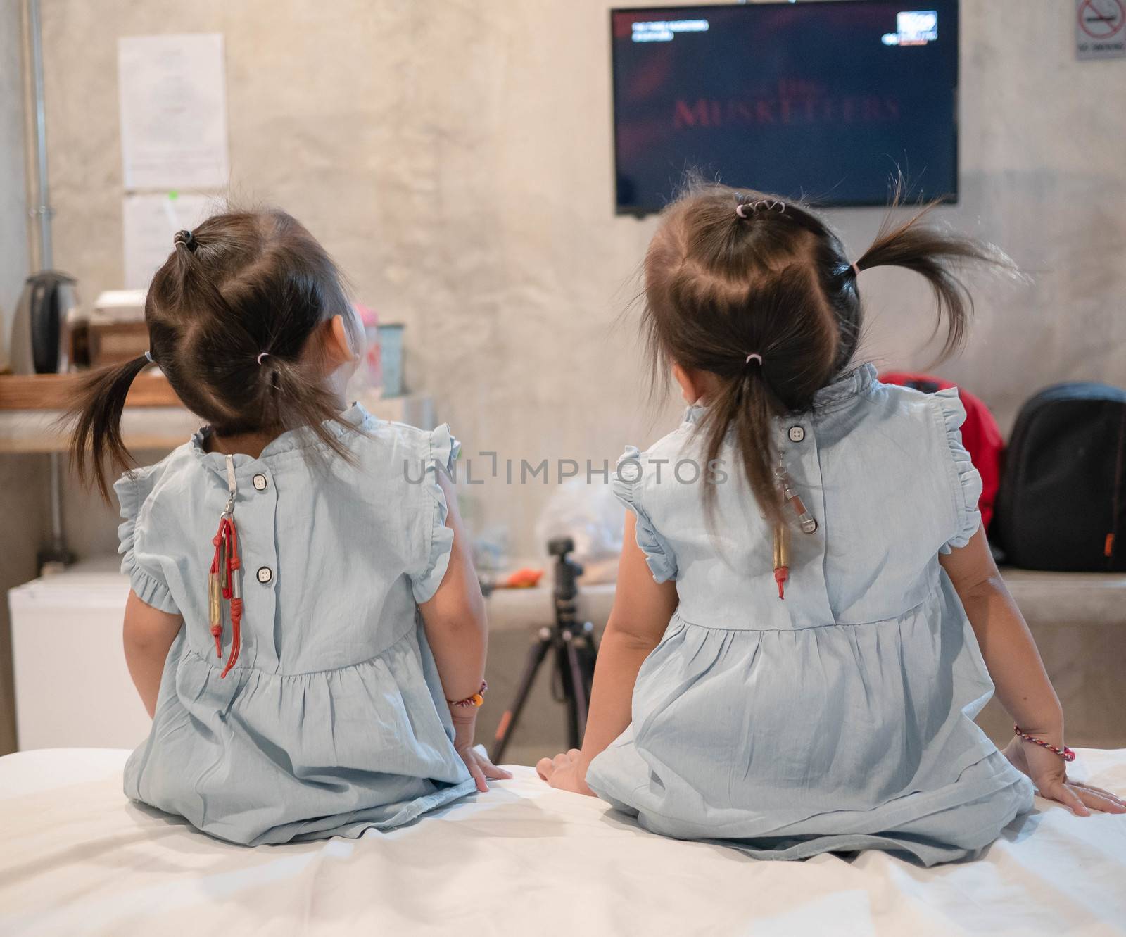 Back view of Asian little child girl sitting on the bed with sister and looking television in the room. by TEERASAK
