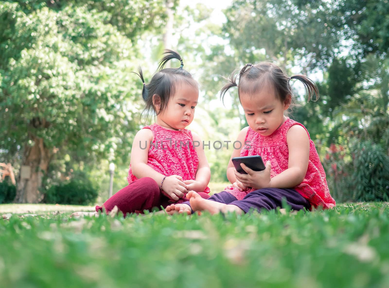 Asian little girl aAsian little girl and sister sitting on the grasses ground in the garden and looking at smartphone happily.nd sister sitting on the grasses ground in th by TEERASAK