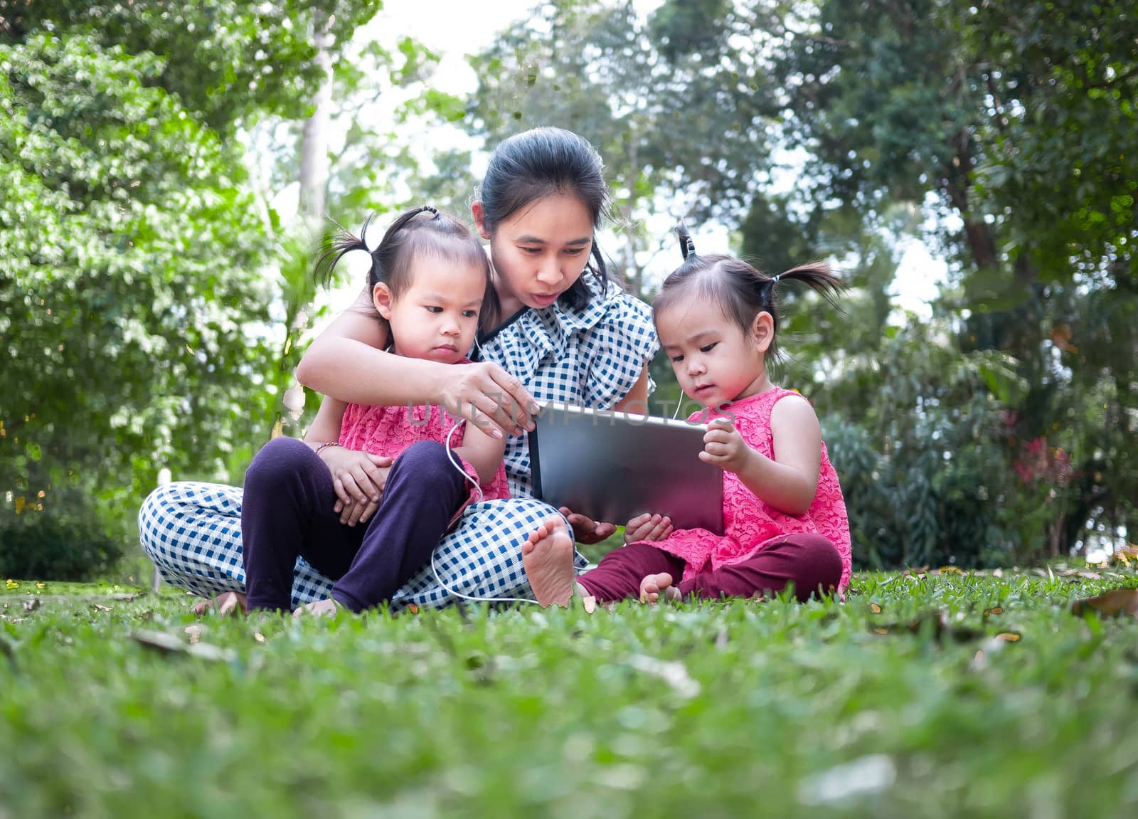 Asian mother and her daughters sitting on the grasses ground in the garden and looking at digital tablet happily. by TEERASAK