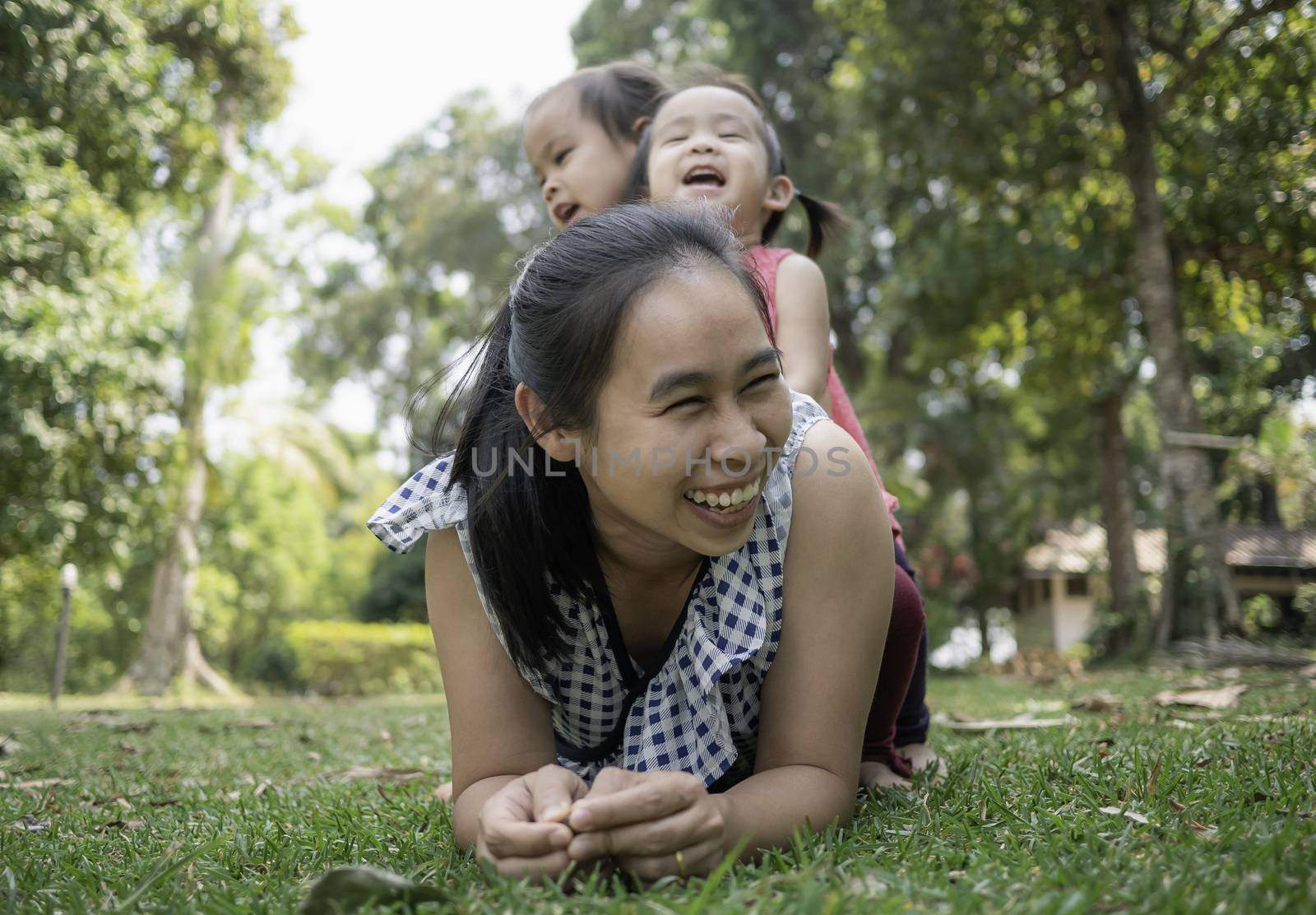 Happy Asian young mother lying on grass and daughters sitting on her back in the park.