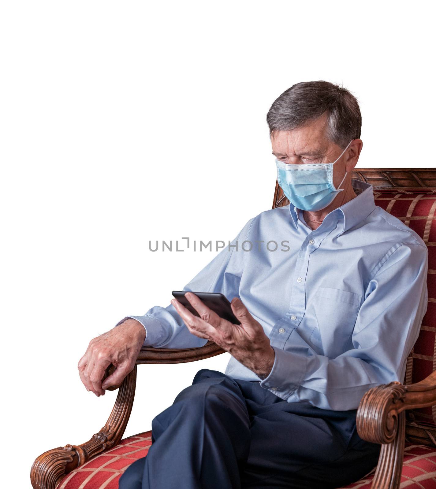 Senior caucasian adult reading from ebook in hand while seated. He is wearing a face mask against coronavirus and cutout against white