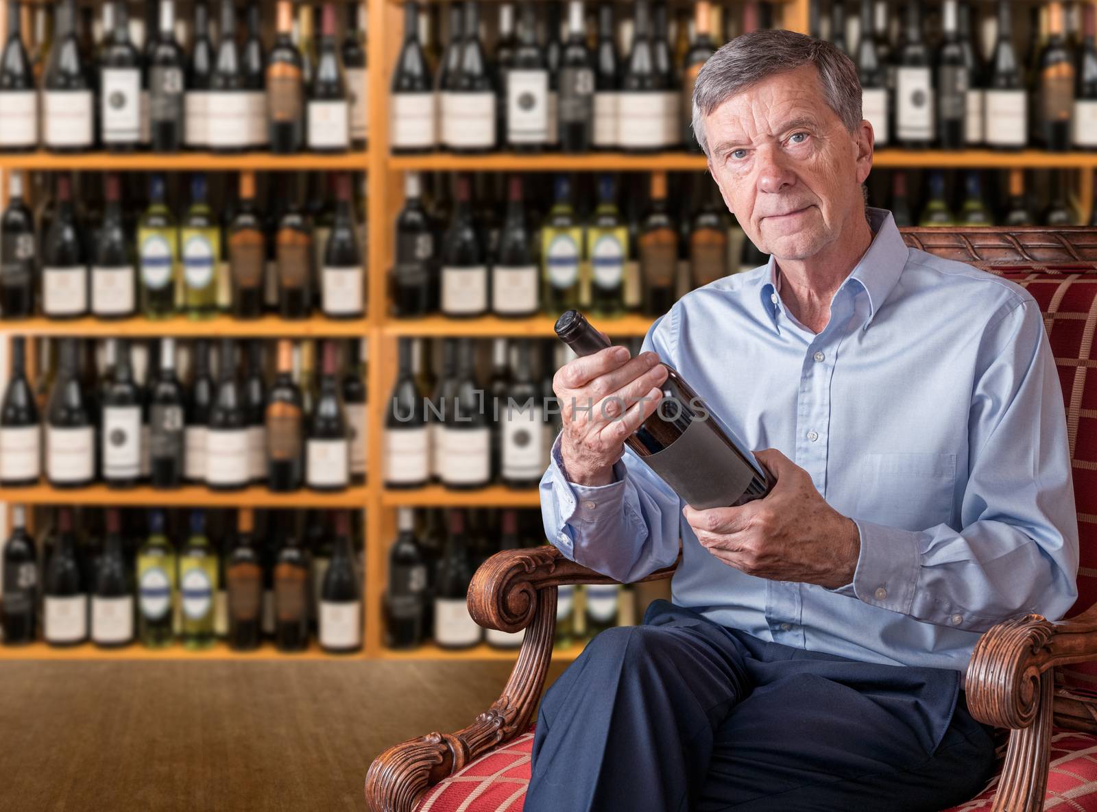 Senior caucasian man examining a wine bottle while seated in front of his shelves by steheap