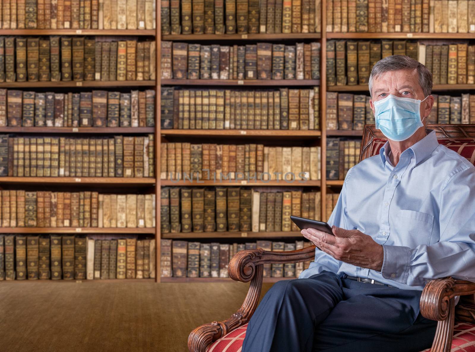 Senior man in face mask against virus reading ebook in antique library by steheap