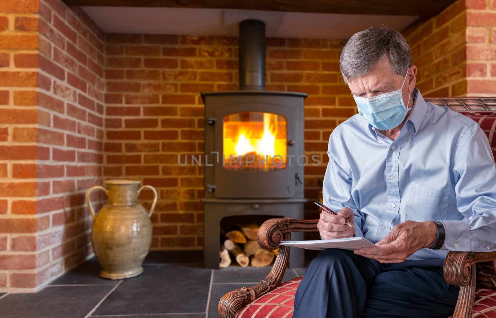 Senior man with face mask against virus checking a document with wood fire in background by steheap