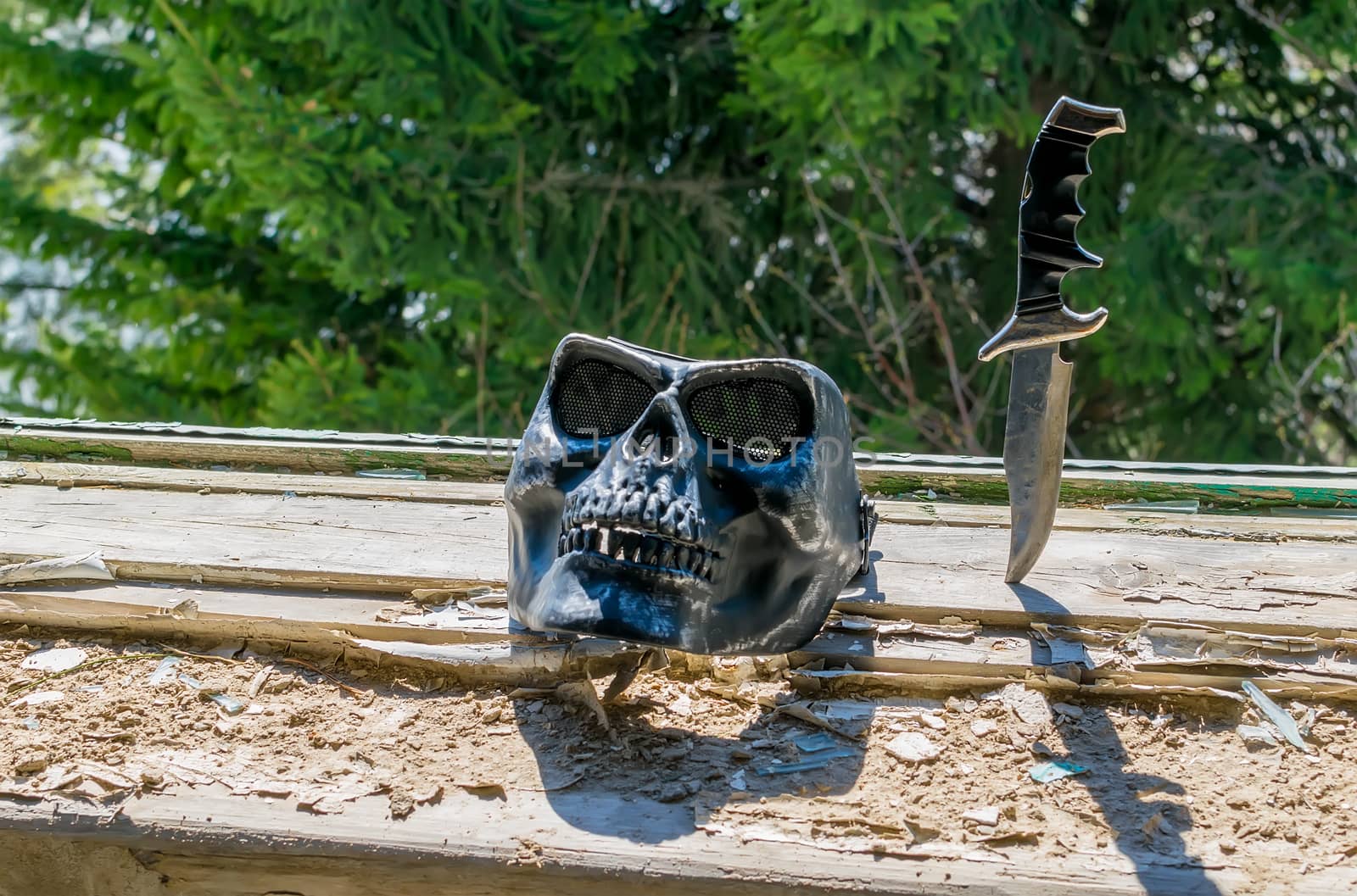 a terrible mask in the form of a skull and an old combat knife stuck in the destroyed windowsill in the window of an abandoned house against the green leaves of a coniferous tree