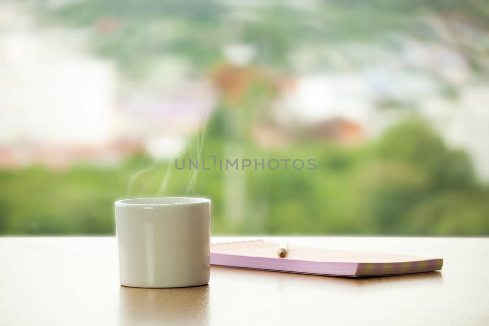 cup of hot coffee with pencil and note book on the table in cafe. cafe window view with green tree for fresh feeling as background. creative and idea work with relax in coffee shop concept