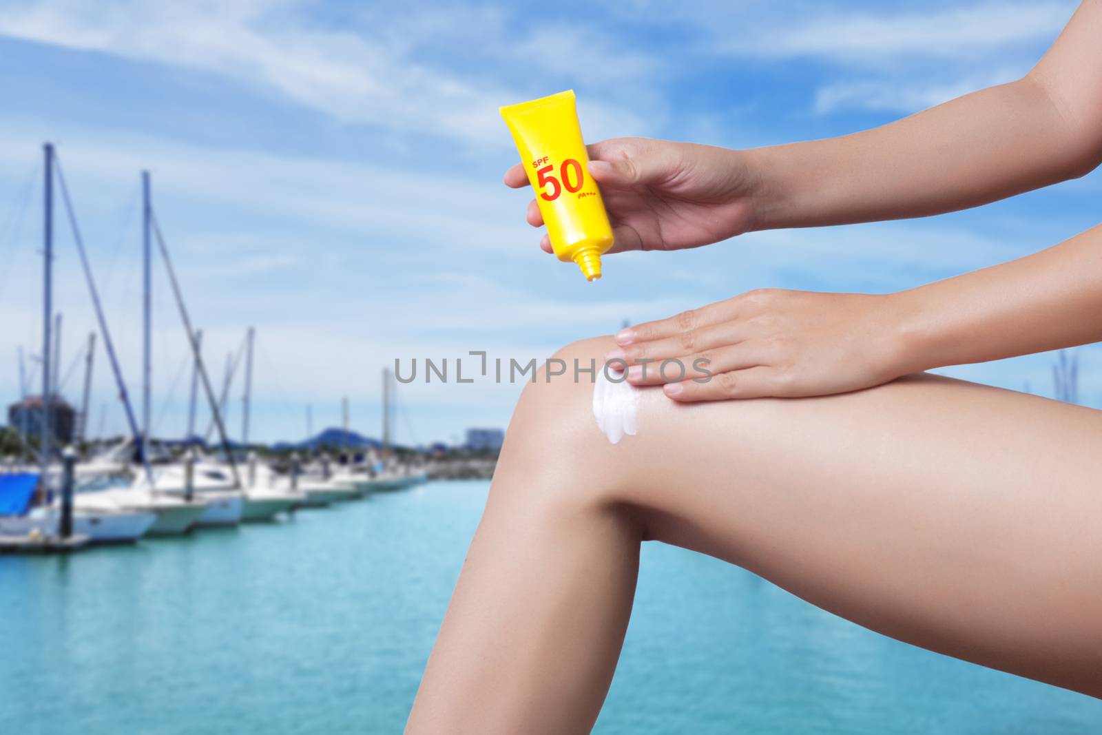 woman applying sunscreen on her leg with blur boats docked at the yacht club background. SPF sunblock protection concept. Travel vacation by asiandelight