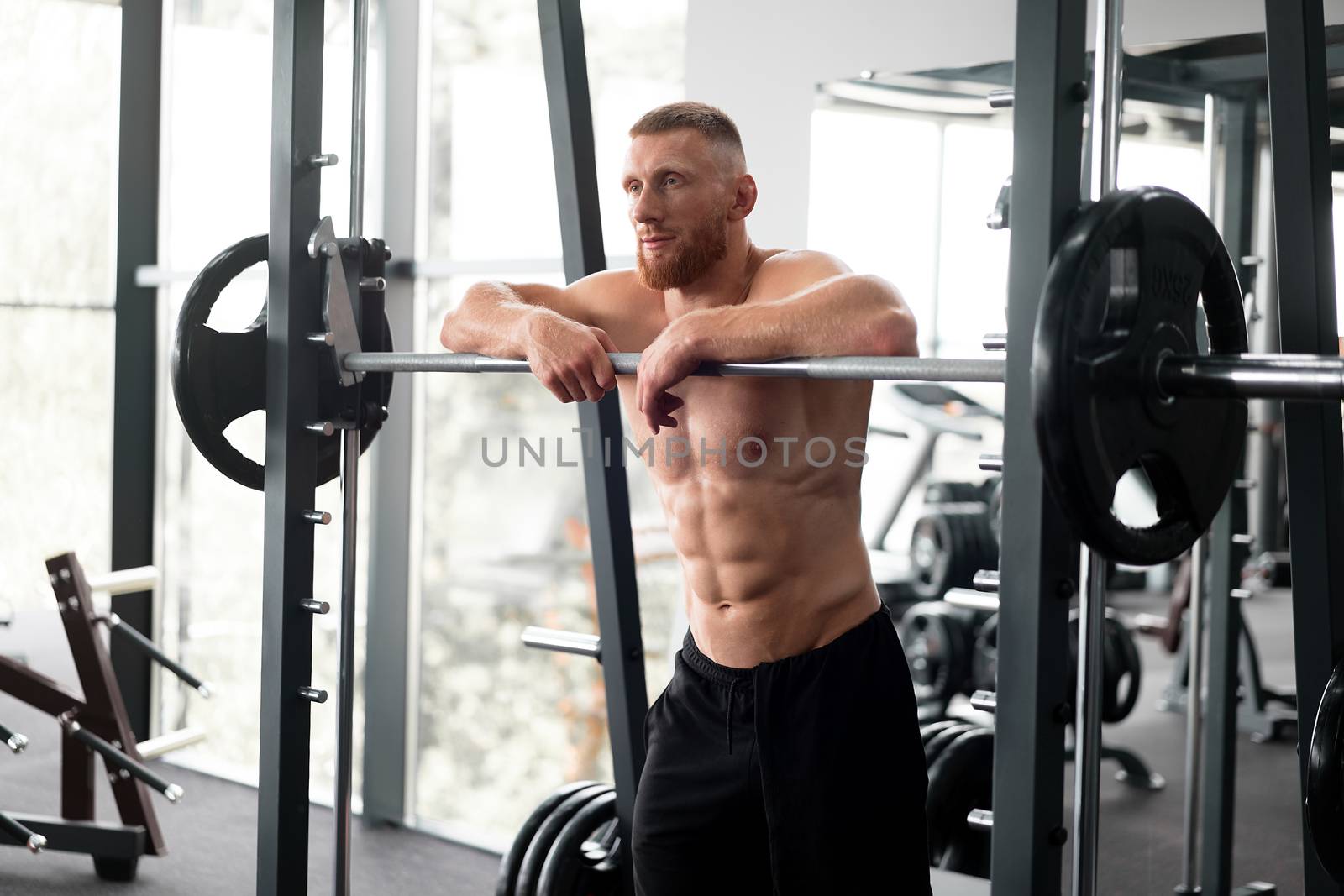 Gym man leaning barbell resting after hard training. Handsome caucasian athlete take break. Sport equipment Sportive guy standing near barbell machine. Perfect muscule male body ideal abs. Naked torso