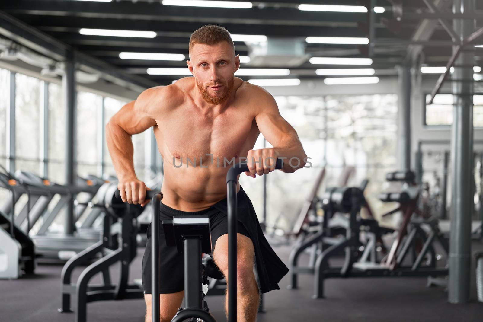 Man exercise bike gym cycling training fitness. Fitness male using air bike cardio workout. Athlete guy naked torso biking indoor gym exercising his legs. Cross functional training.