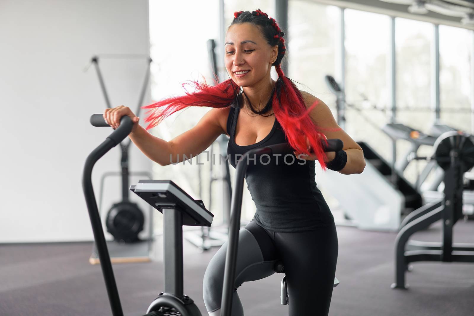 Woman exercise bike gym cycling training fitness. Fitness female using air bike cardio workout. Athlete girl sportswear biking indoor gym exercising his legs. Cross functional training.