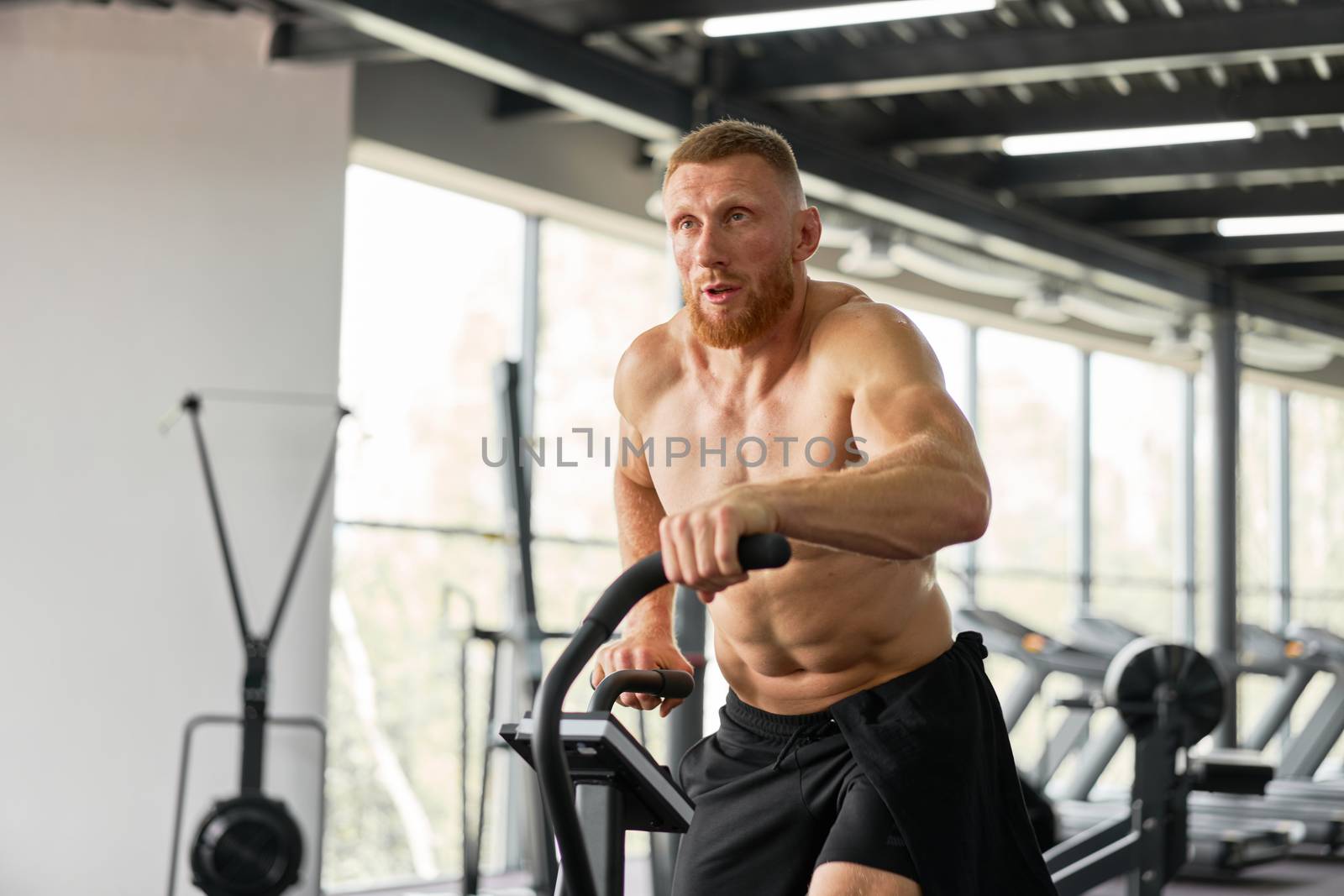 Man exercise bike gym cycling training fitness. Fitness male using air bike cardio workout. Athlete guy naked torso biking indoor gym exercising his legs. Cross functional training.