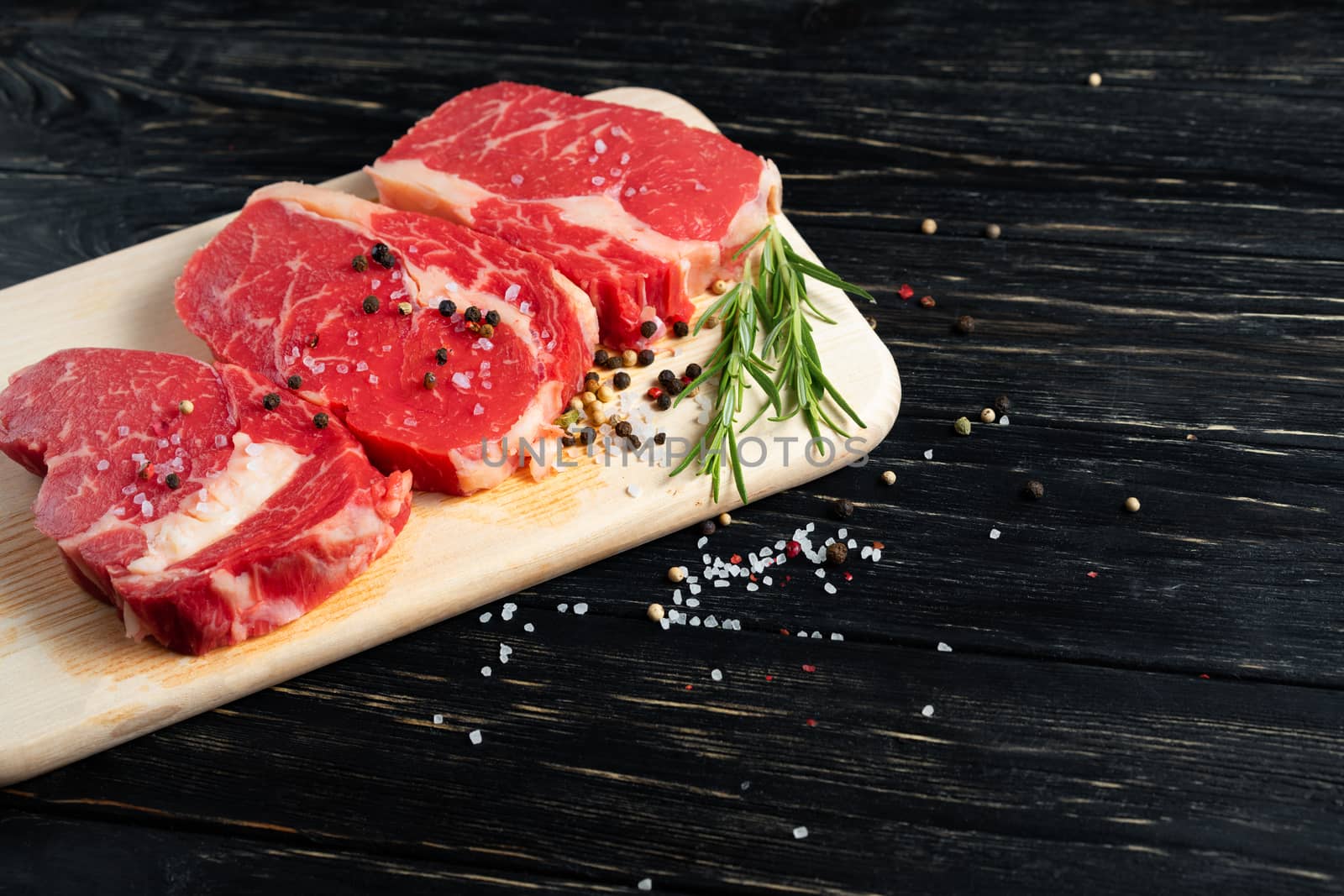 Three pieces of juicy raw beef with rosemary on a cutting board on a black wooden table background. Meat for barbecue or grill sprinkled with pepper and salt seasoning