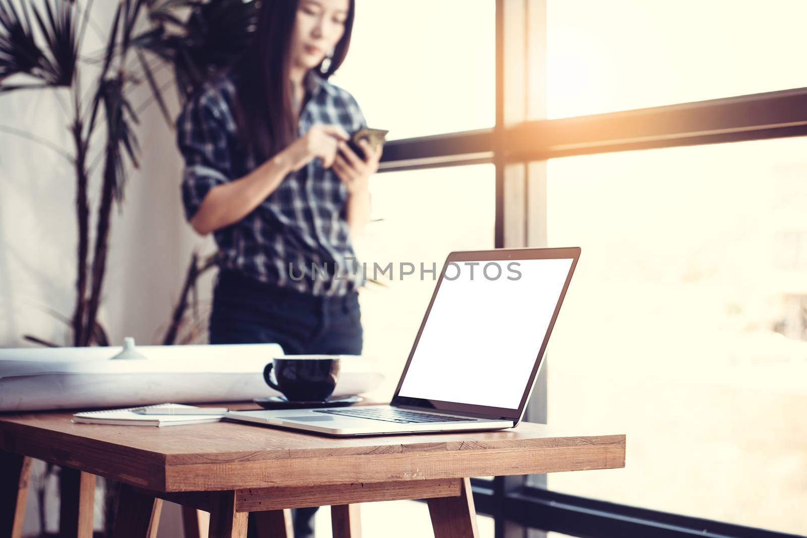 Technology in workplace concept. Young Asian business woman in modern office using the phone for work. Blank white screen laptop and blueprint on desk. by asiandelight