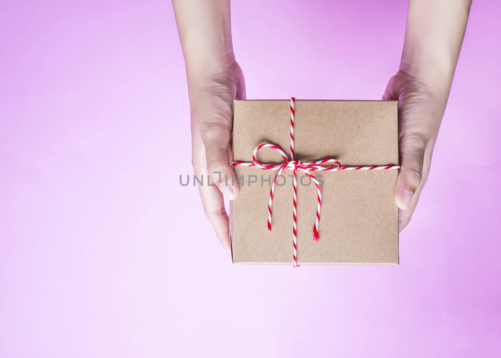 Female hands holding a simple craft paper gift box wrapped with colorful rope with as a present for Christmas, new year, valentine day or anniversary on pink background, top view by asiandelight