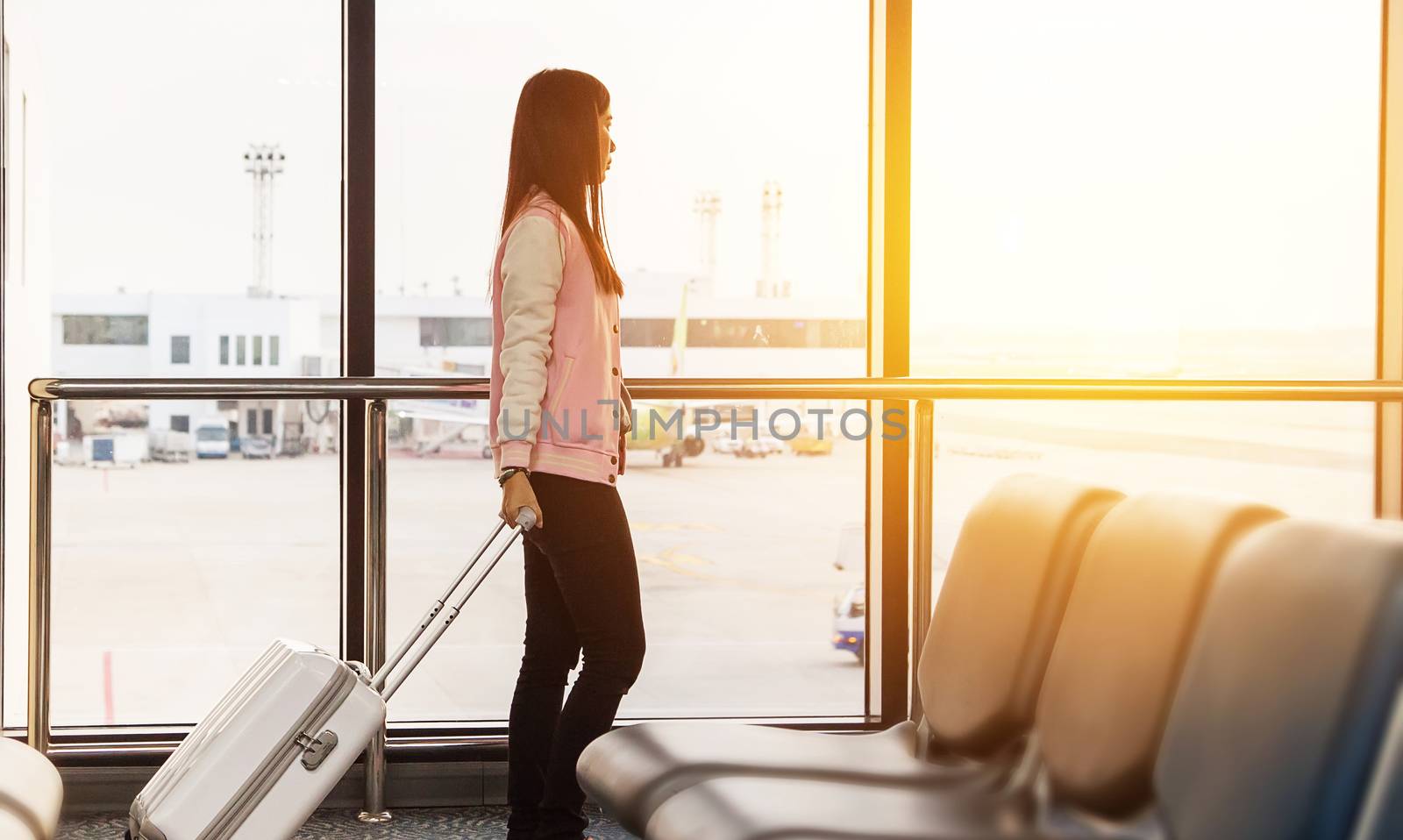 Travel lifestyle concept. Travel tourist woman waiting at boarding gate terminal before departure with luggage looking at airplanes with sunrise view at airport window for morning flight.